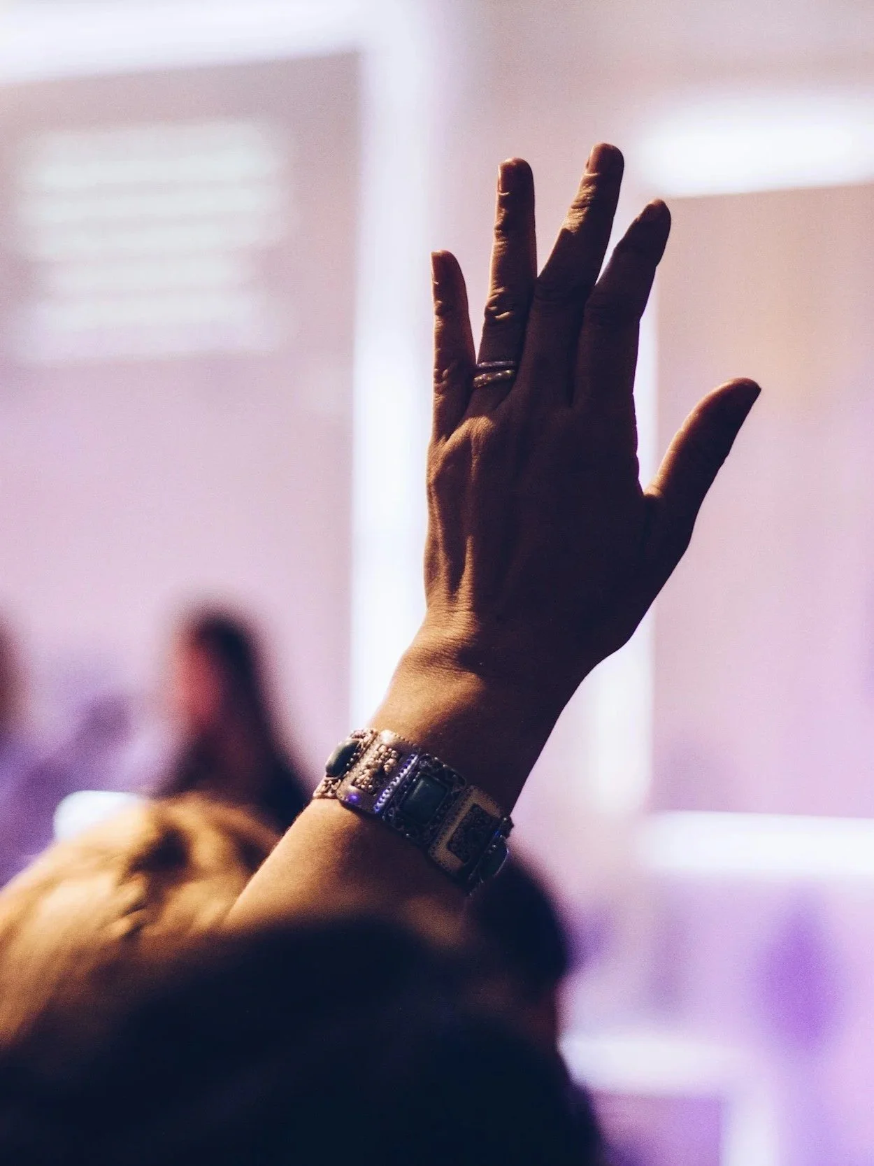 A person raising their hand with fingers spread apart, wearing a bracelet and a ring, in a dimly lit environment.