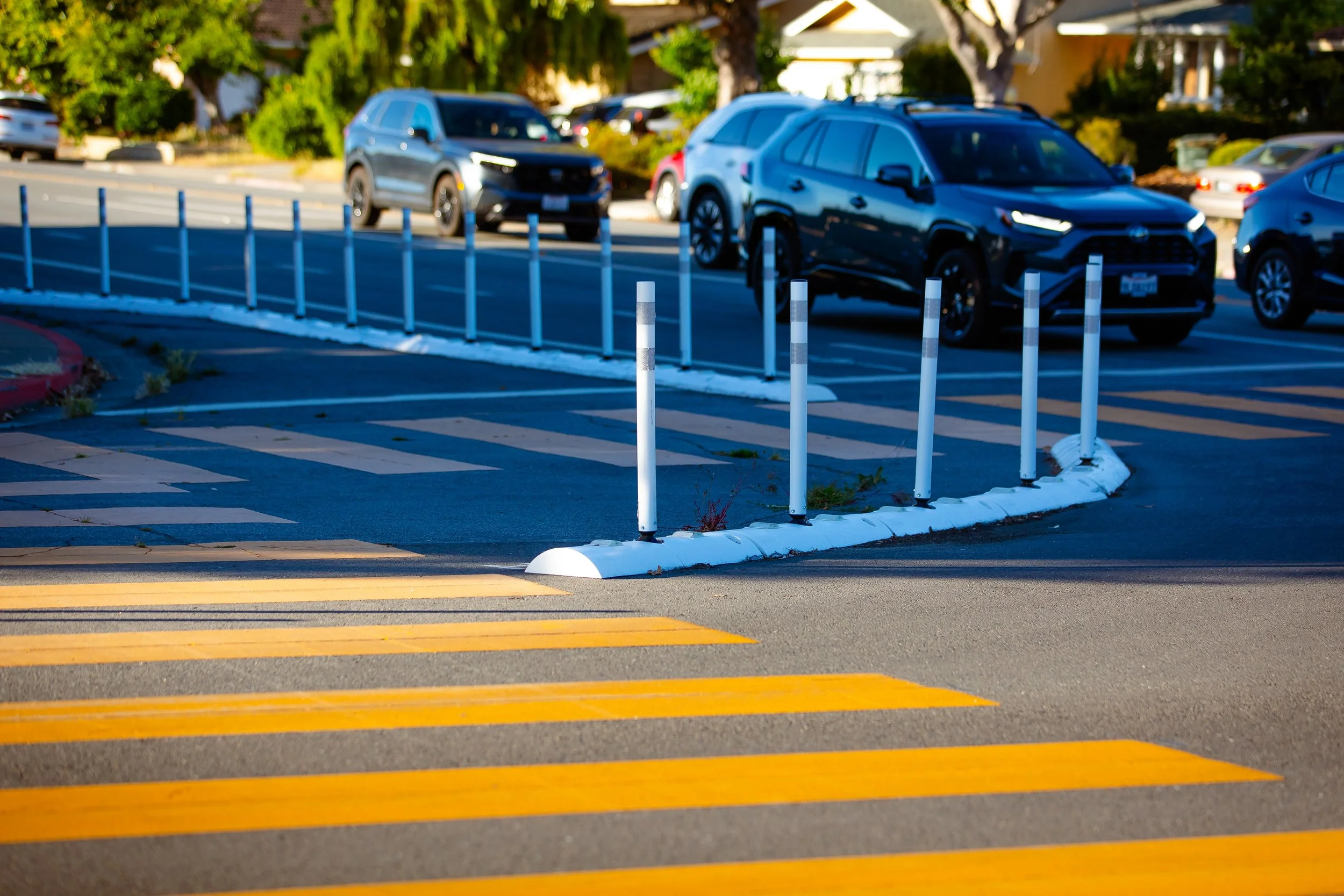A protected bike lane on a street in San Jose, CA, near a parking lot with several parked cars, a black SUV in the foreground, and a row of white posts separating the parking area from the crosswalk with yellow stripes.