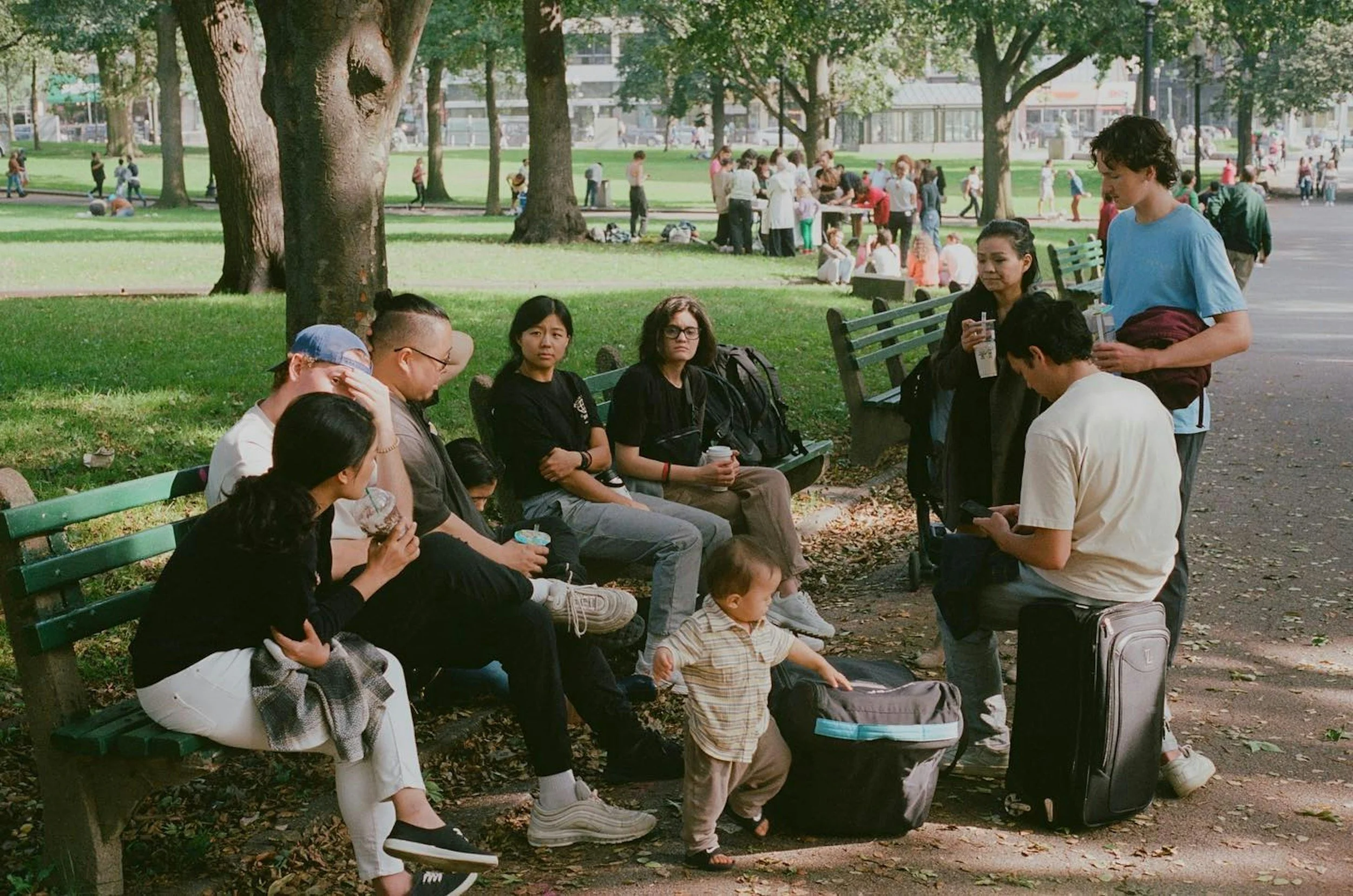 A group of community members is sitting on benches and standing in a park with trees and grass. Some are drinking and looking at their phones, while a small child walks near a backpack.