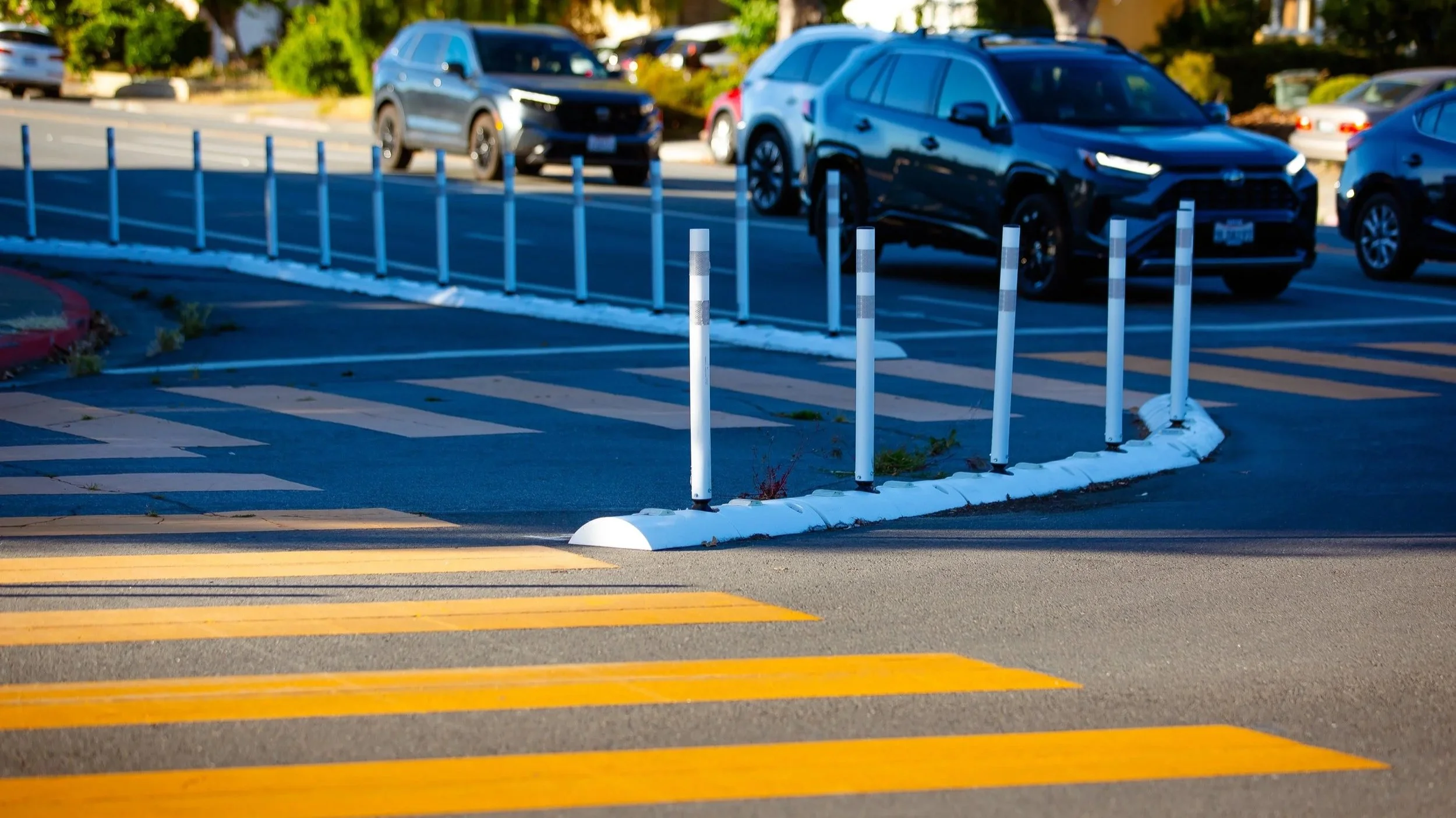 A protected bike lane on a street in San Jose, CA, near a parking lot with several parked cars, a black SUV in the foreground, and a row of white posts separating the parking area from the crosswalk with yellow stripes.