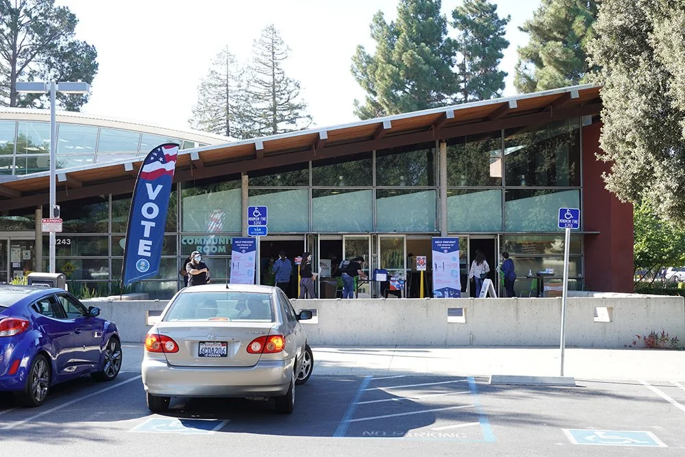 People at a public library in San Jose, CA. The library has a voting polling station with a large 'VOTE' banner outside, and cars parked in a lot in front.