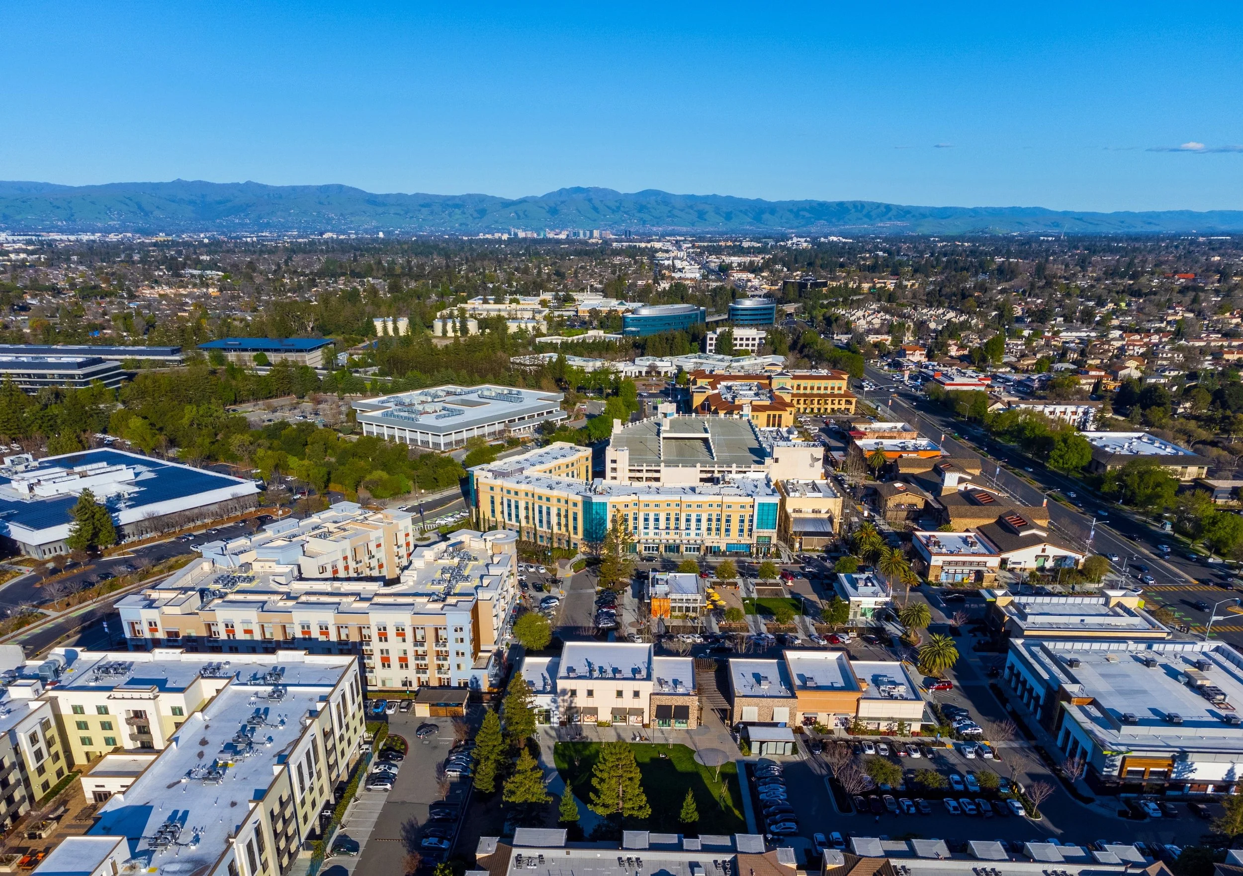 An aerial view of a San Jose, CA cityscape with various commercial and residential buildings surrounded by trees and streets, with mountains in the background under a clear blue sky.