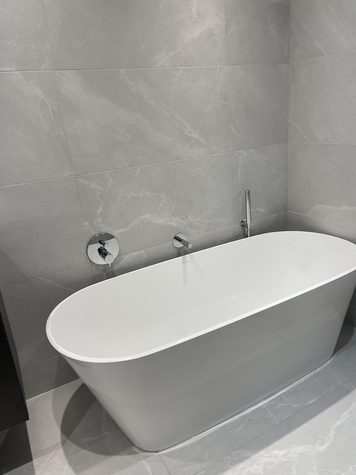 Modern white freestanding bathtub in a bathroom with grey marble tiled walls and floor.