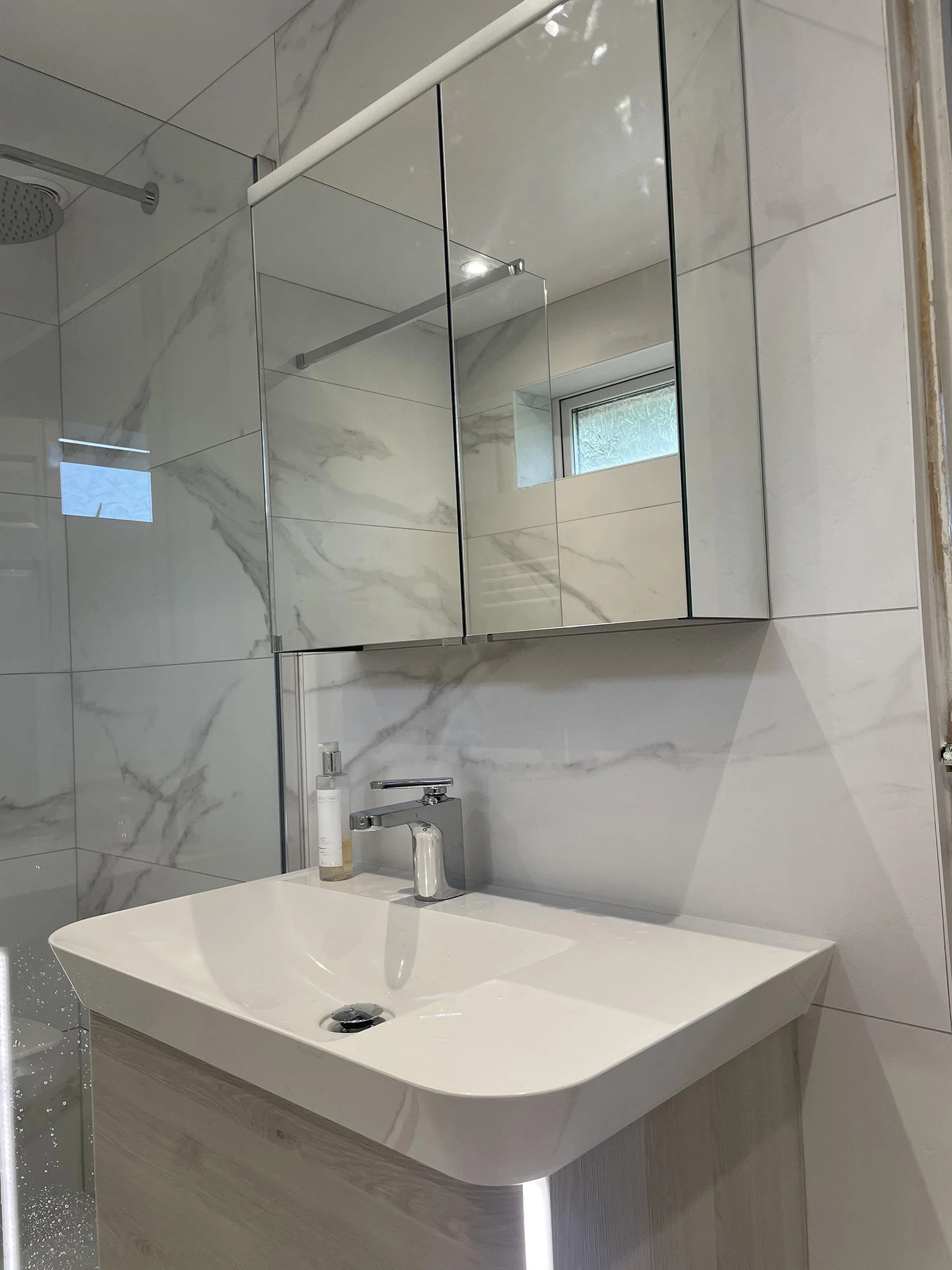 Modern bathroom with a white sink, a wall-mounted faucet, a mirrored cabinet, and a small window with frosted glass, all featuring a marble tile wall.