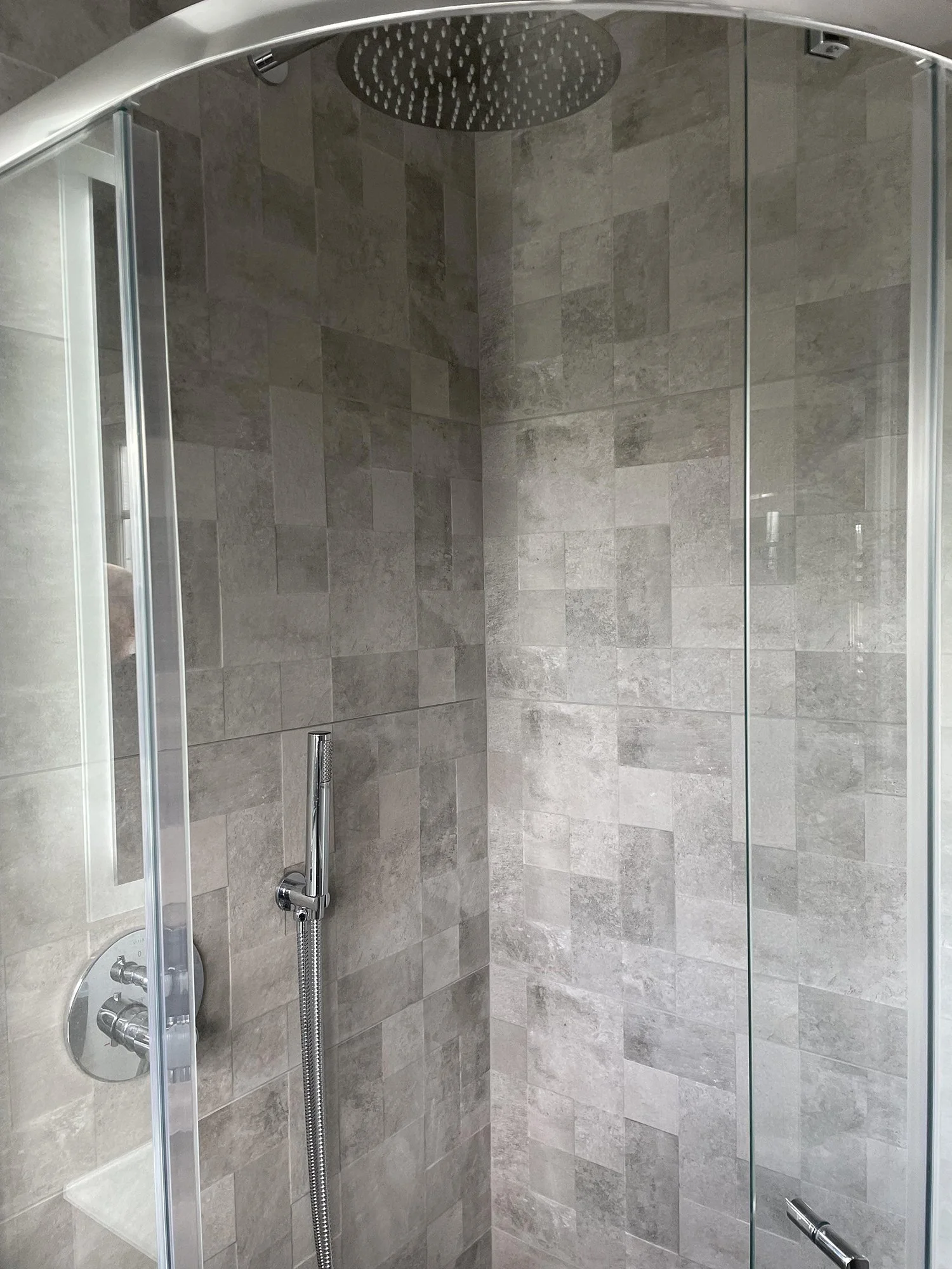 Modern shower with grey ceramic tiles, a rainfall showerhead, a handheld showerhead, and glass enclosure.