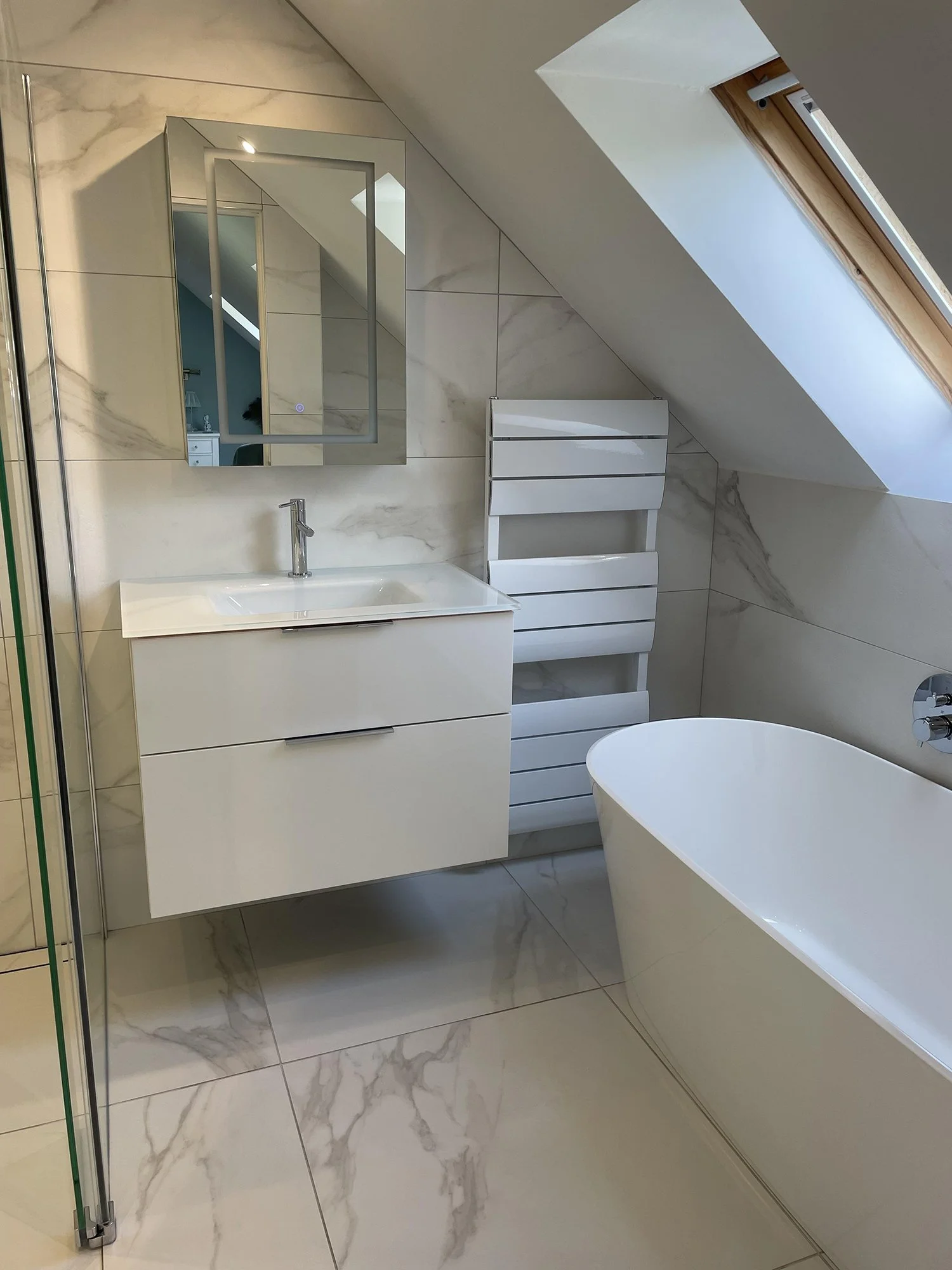 A modern bathroom with a white vanity, a rectangular mirror, a bathtub, a sloped ceiling with a skylight, a towel radiator, and marble-patterned tiles.