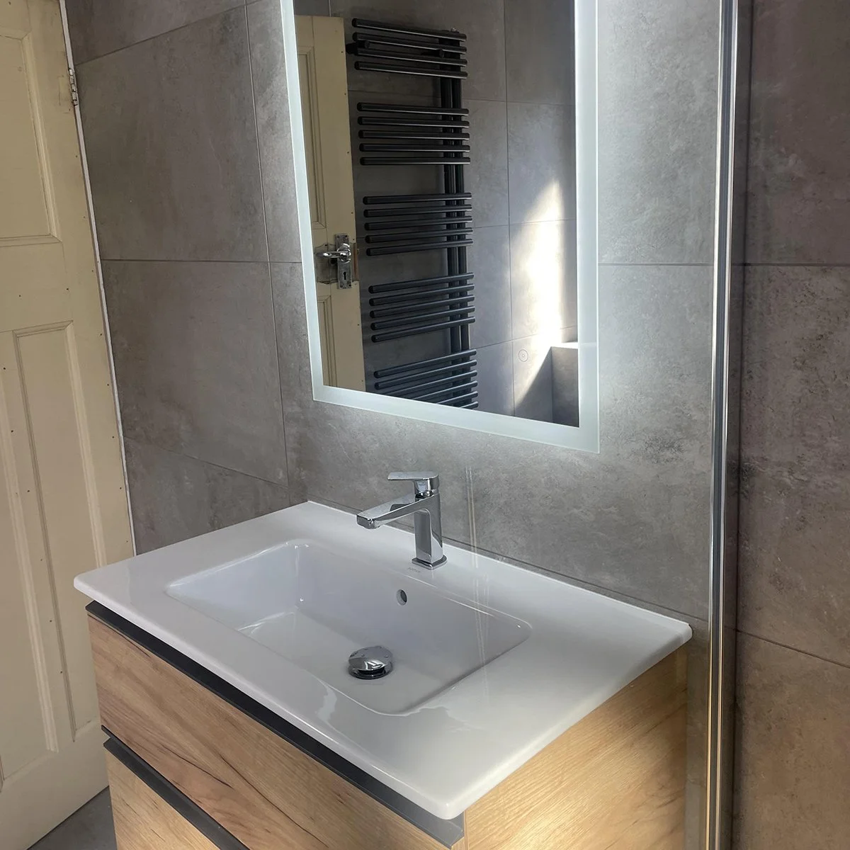 A modern bathroom sink with a white ceramic basin, a silver faucet, and a wooden cabinet below. A large mirror is mounted on the wall above the sink, reflecting black heated towel radiators on the opposite wall. The walls are tiled in grey.