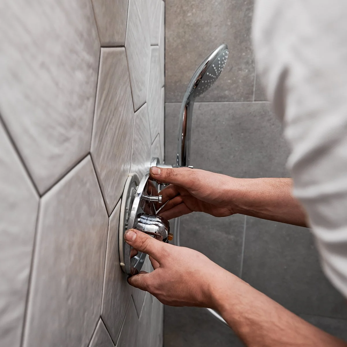 A person is adjusting a shower faucet in a bathroom with grey tiled walls.