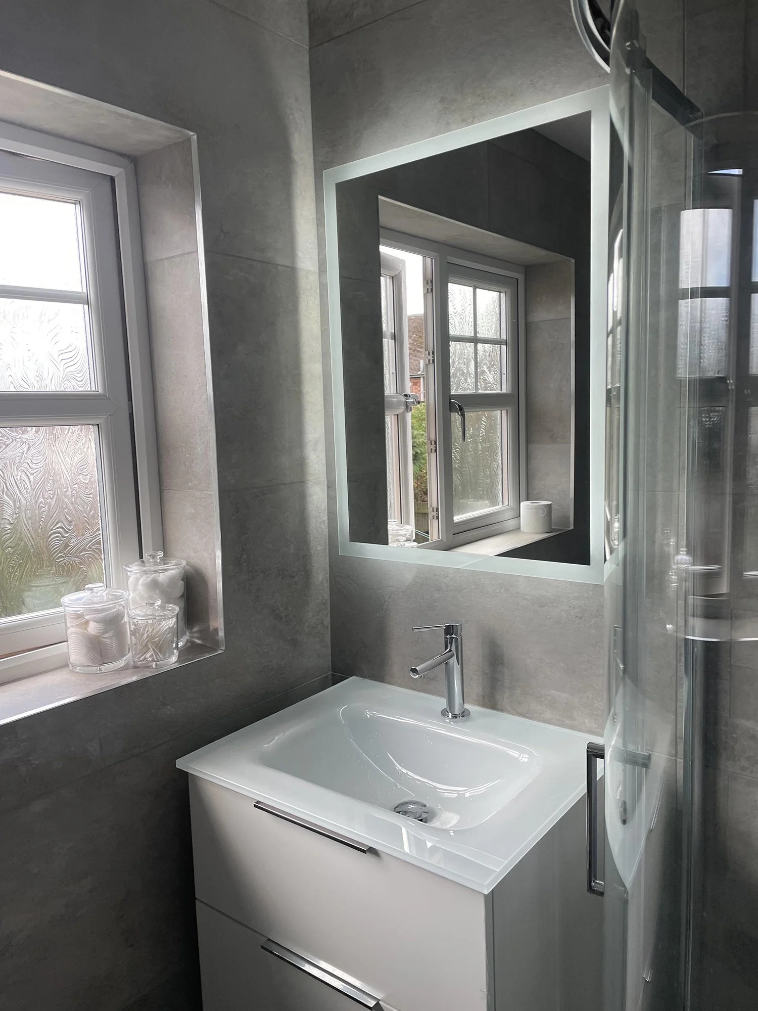 A modern bathroom with a white vanity, a small sink, a wall mirror, and a window with frosted glass. The walls are tiled in grey.