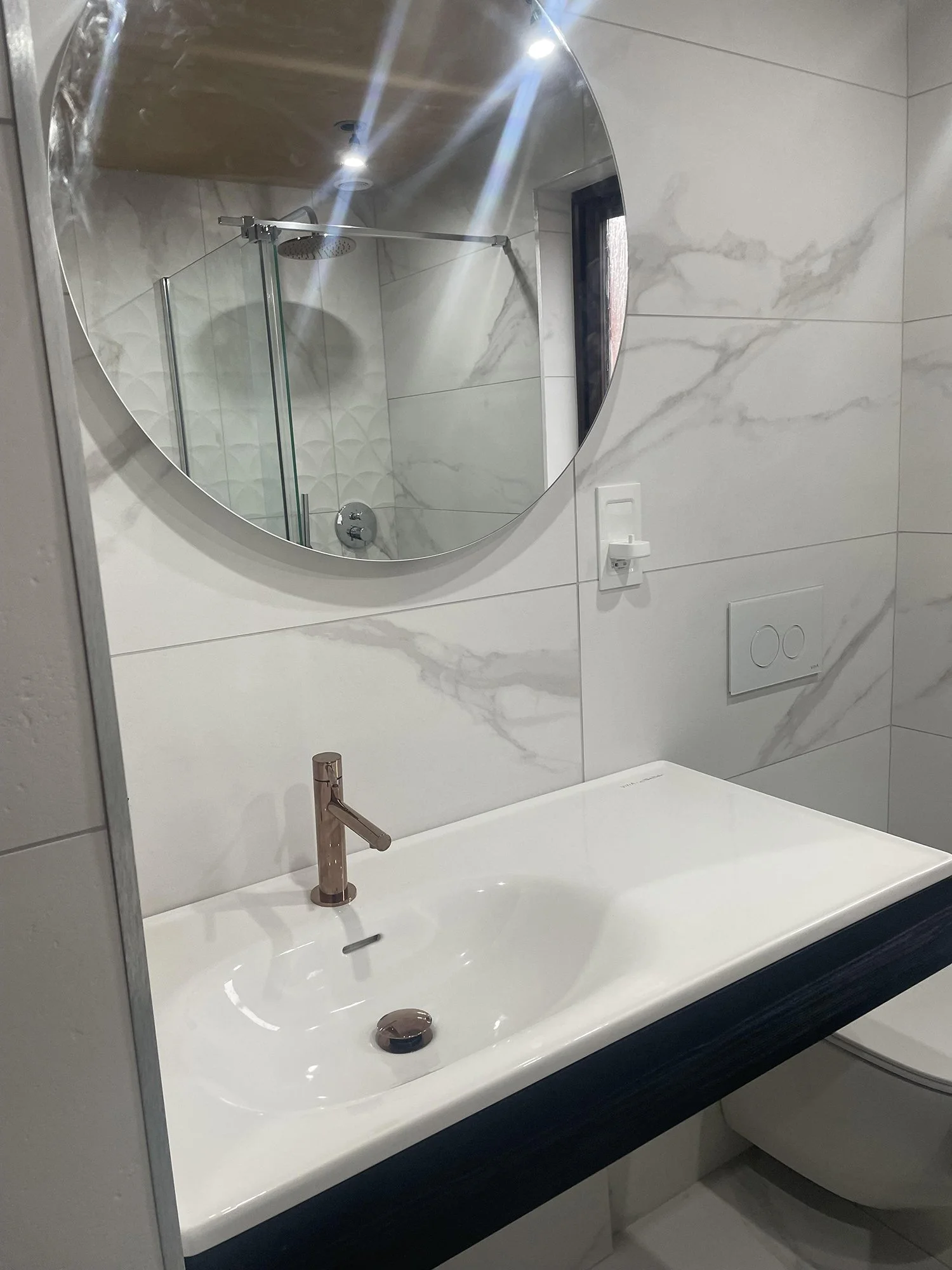 Modern bathroom with a white sink, rose gold faucet, round mirror, and a shower area with glass door and rain shower head reflected in the mirror.