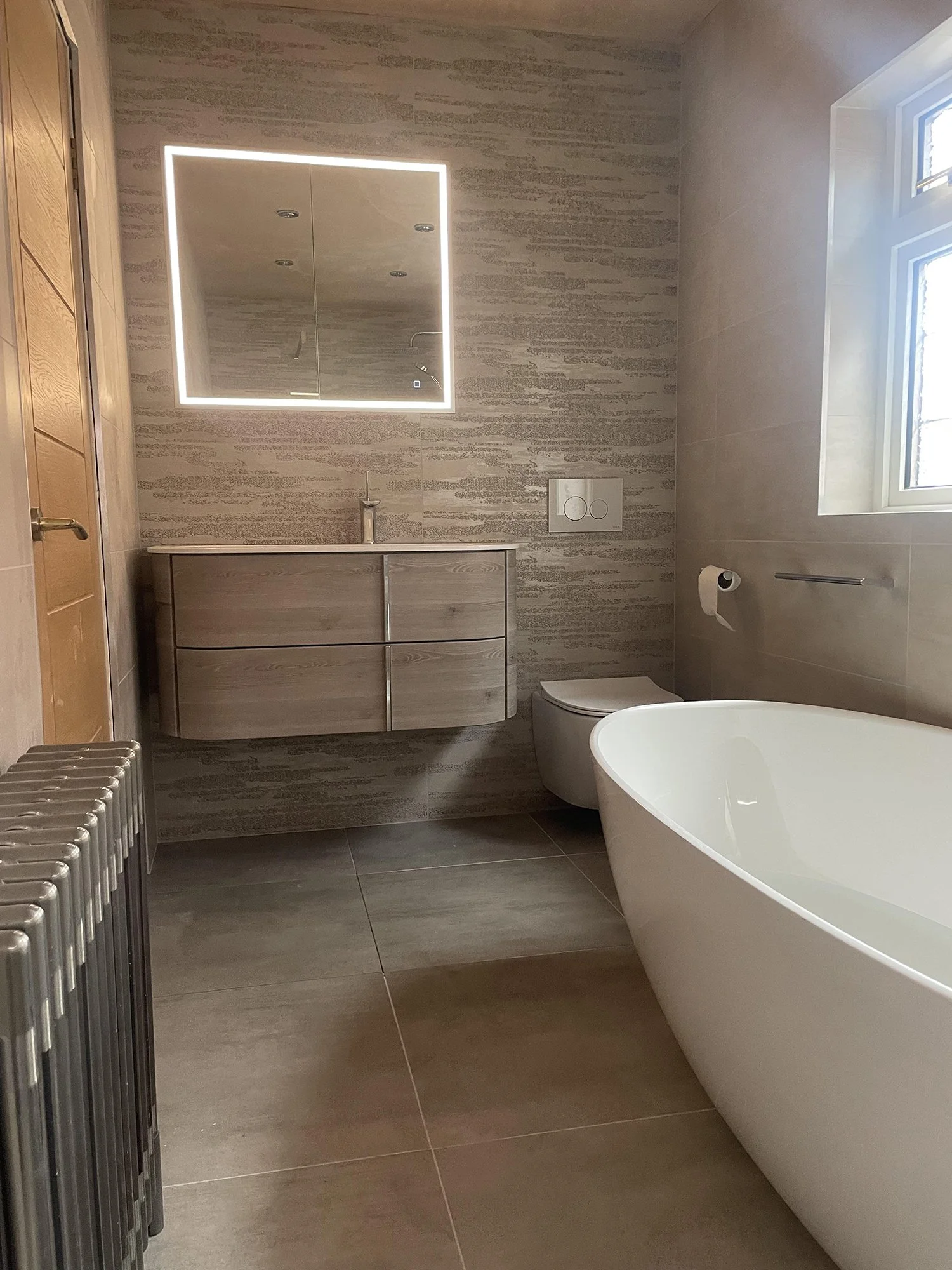 Modern bathroom with bathtub, toilet, wooden vanity with sink, illuminated mirror, and a window letting in natural light.