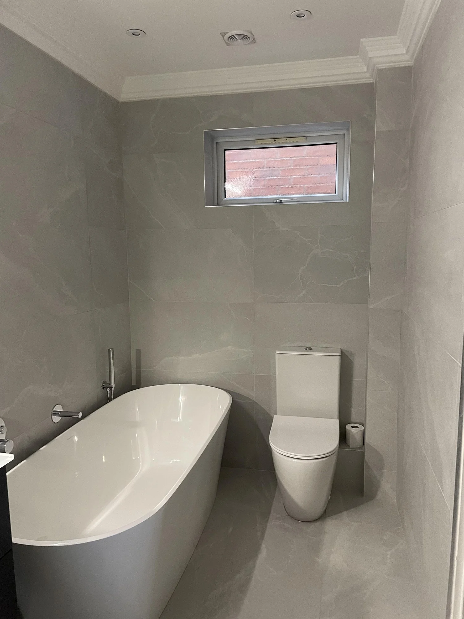 Modern bathroom with grey marble walls and floors, a white bathtub, a white toilet, a small window, and a roll of toilet paper.