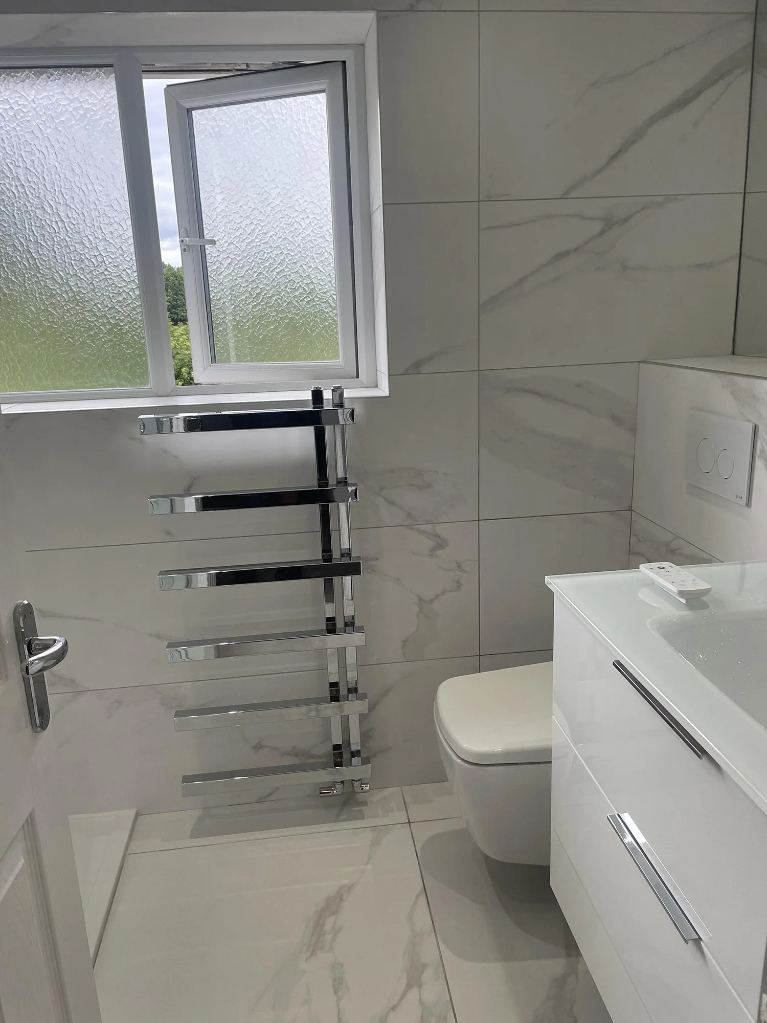 A modern bathroom with white marble walls and floor, a frosted window, a wall-mounted toilet, a white vanity with two drawers, a remote control on top, and a metallic heated towel rail next to the window.
