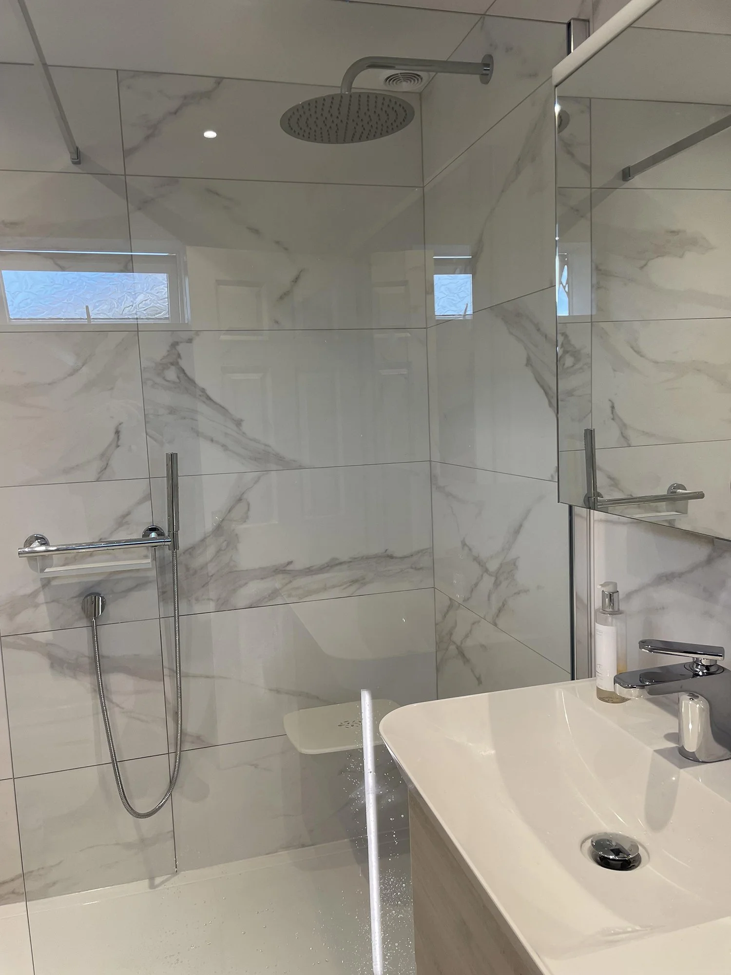 Modern bathroom with a rainfall showerhead, marble-tiled walls, a glass shower enclosure, a small bathroom mirror, and a white sink with a faucet and soap dispenser.