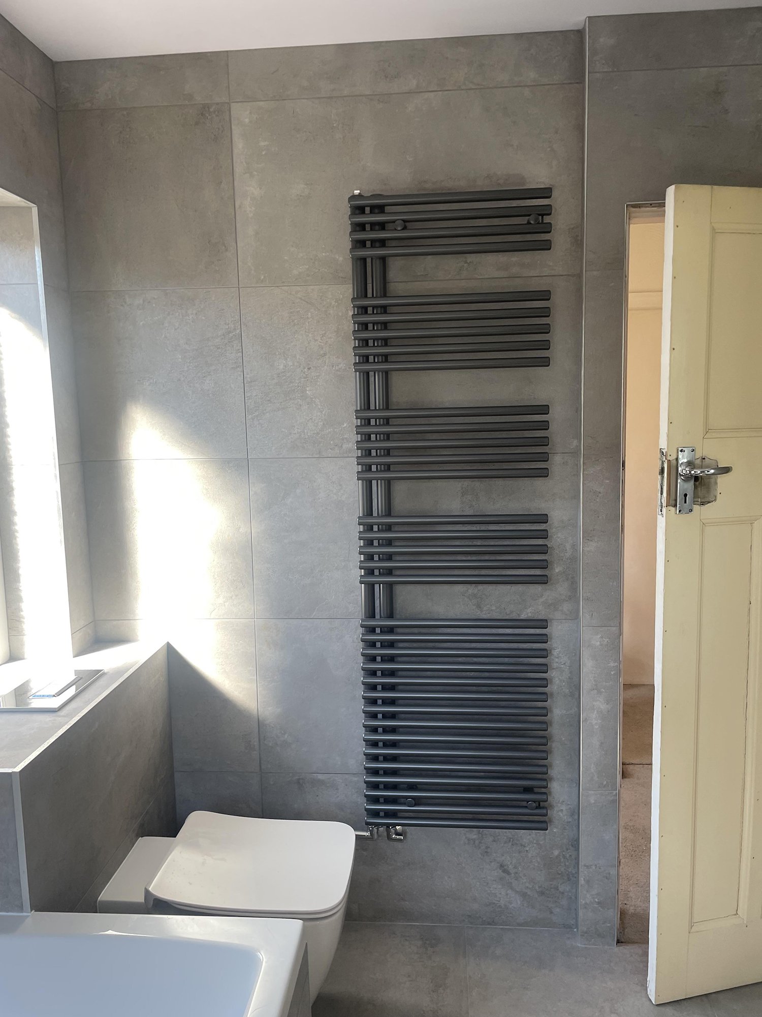 Grey tiled bathroom with a window, white toilet, and a black heated towel rail on the wall.
