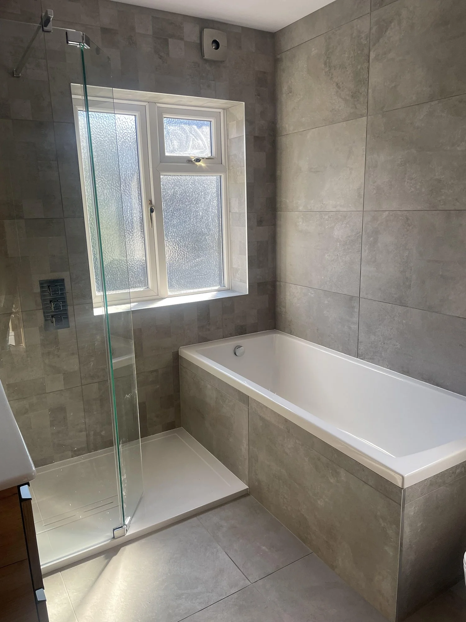 Modern bathroom with a large bathtub, walk-in shower with glass door, grey tiled walls and floor, and a frosted window.