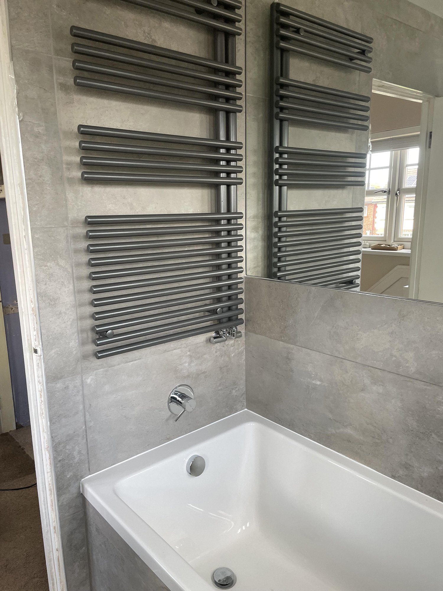 Modern bathroom with a white bathtub, grey tiled walls, a large mirror, and two heated towel rails mounted on the wall above the tub.