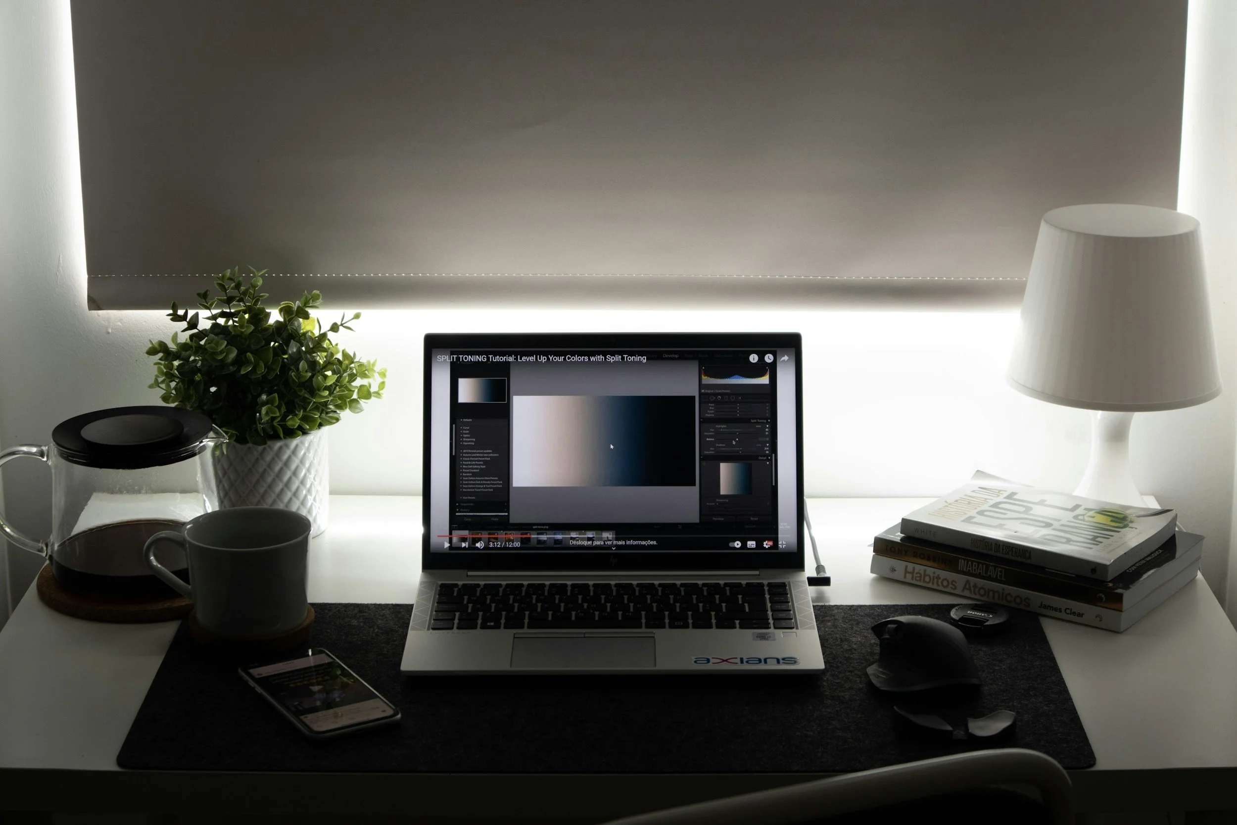 A workspace with a laptop displaying a video tutorial on split toning, a potted plant, a teapot with a cup, a smartphone, a computer mouse, a stack of books, and a white table lamp, all on a white desk with a black desk mat.