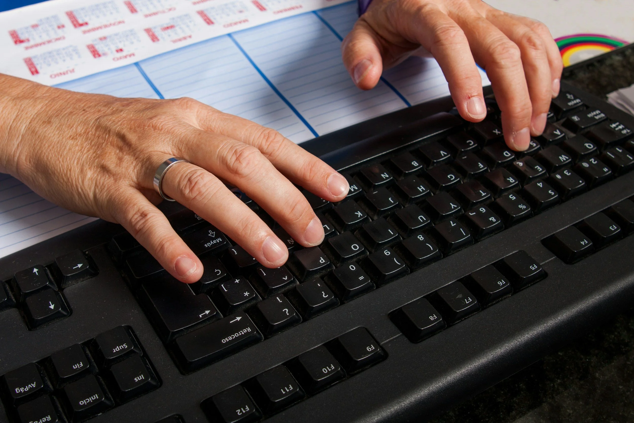A person's hands typing on a black computer keyboard. The person is wearing a silver ring on their left ring finger and has short, clean nails. There are printed papers and a colorful object in the background.