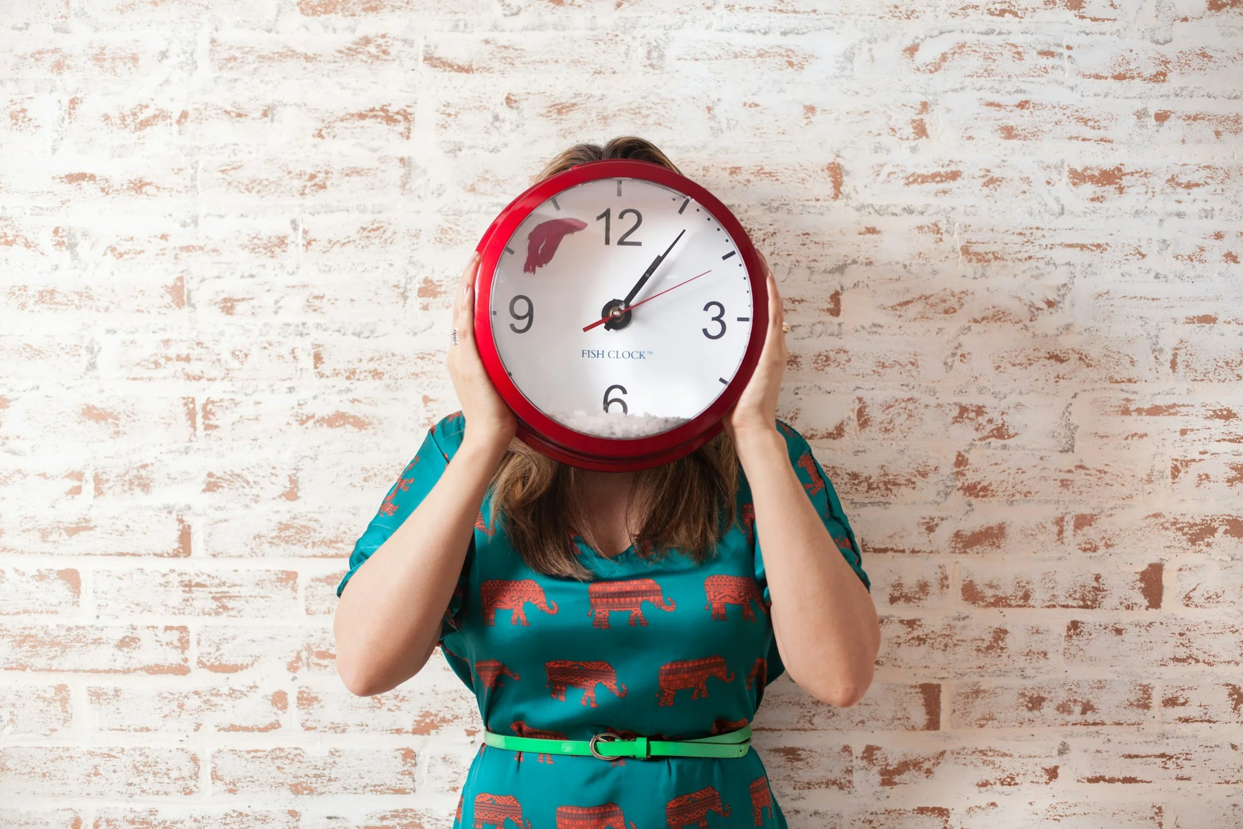 A woman holding a large red fish-shaped clock in front of her face, with a brick wall in the background.