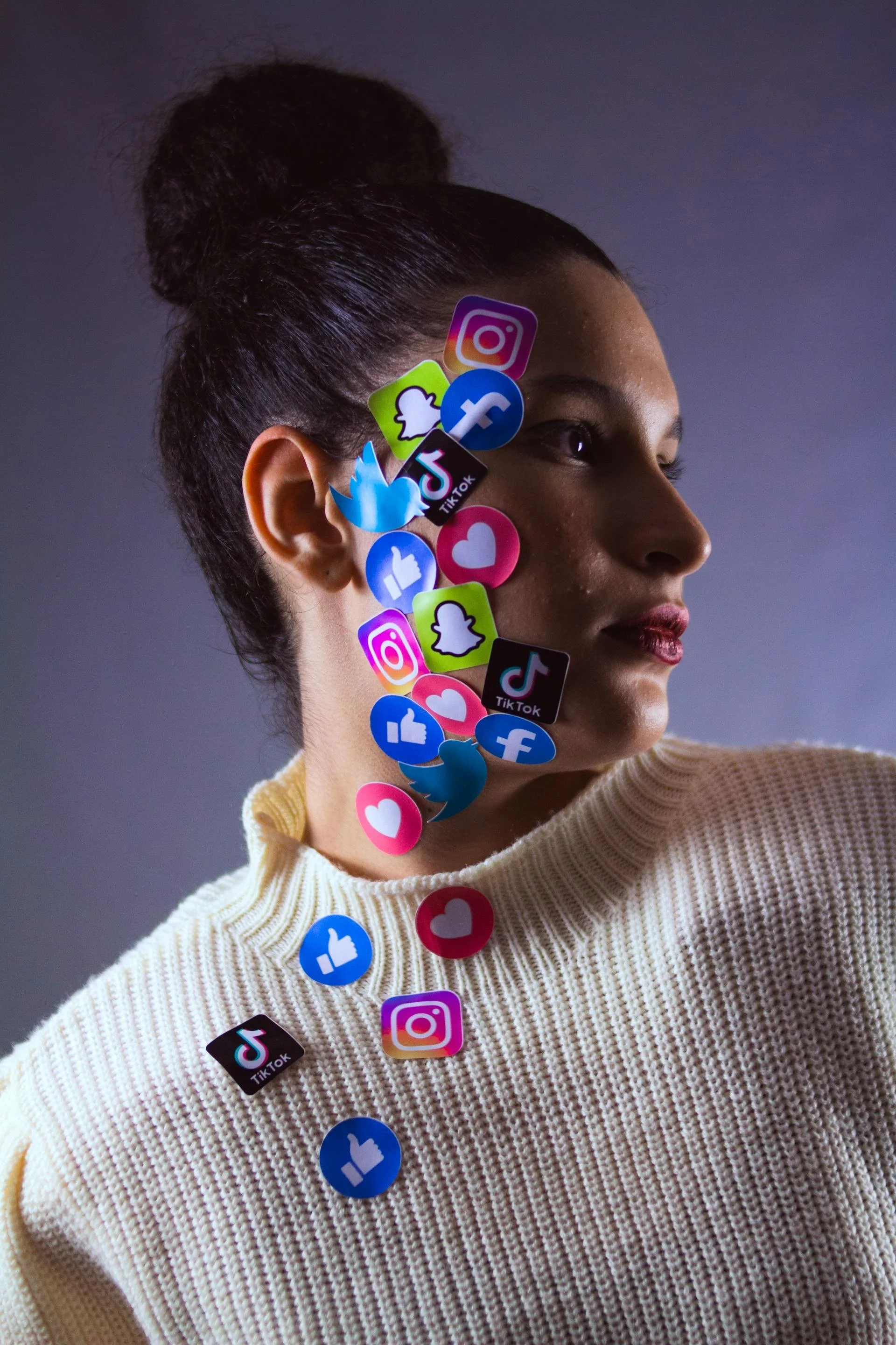 A woman with dark hair in a bun, wearing a cream-colored sweater, with various social media icons such as Instagram, TikTok, Snapchat, Facebook, Twitter, and like or love symbols placed on and around her face and shoulder.