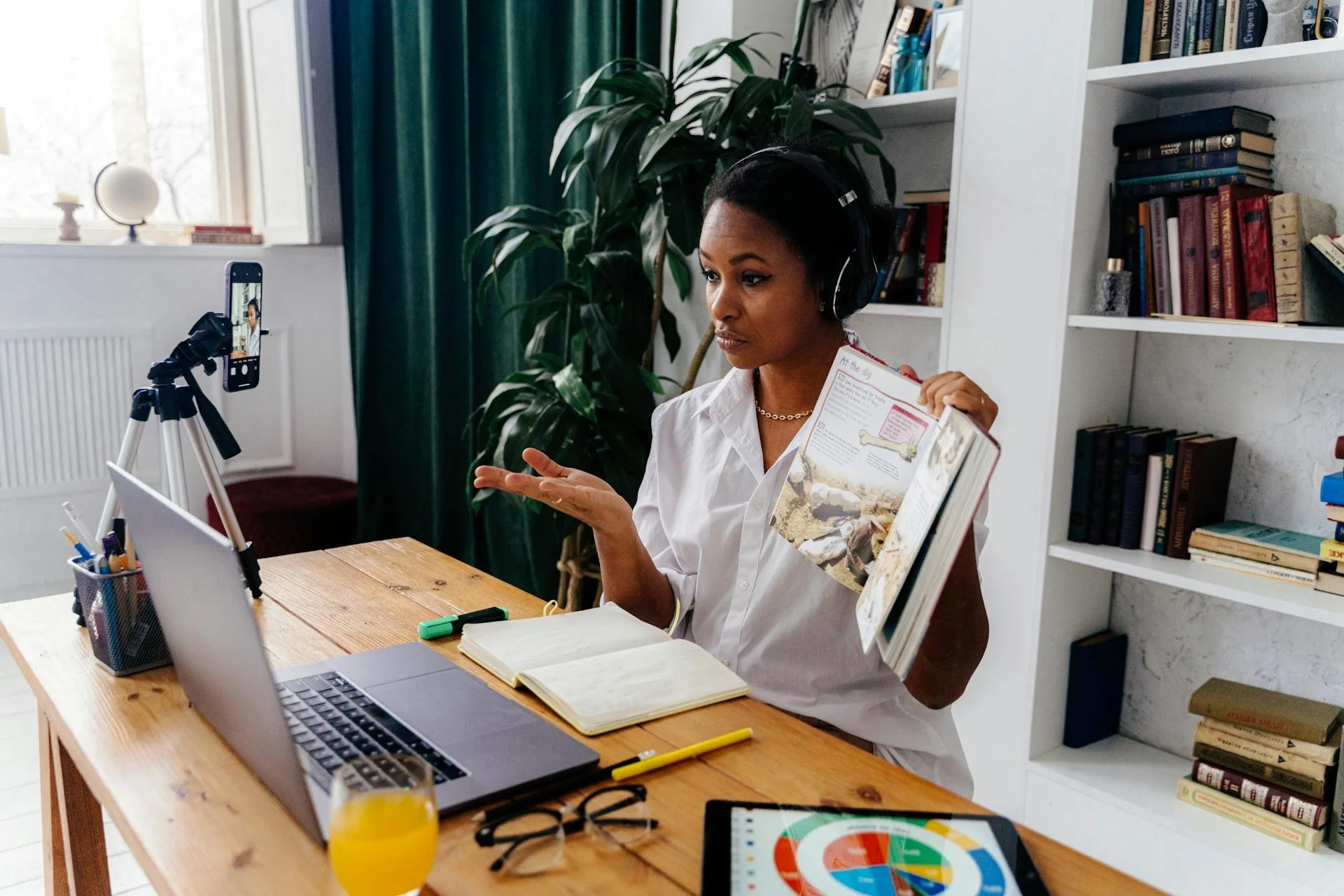 A woman wearing headphones is sitting at a wooden desk with a laptop, open notebook, and various stationery items. She is holding a colorful magazine or textbook in her left hand and gesturing with her right hand. A glass of orange juice, a pair of glasses, and a chart are on the desk. A camera on a tripod is recording her, and behind her is a bookshelf filled with books and a large green plant.