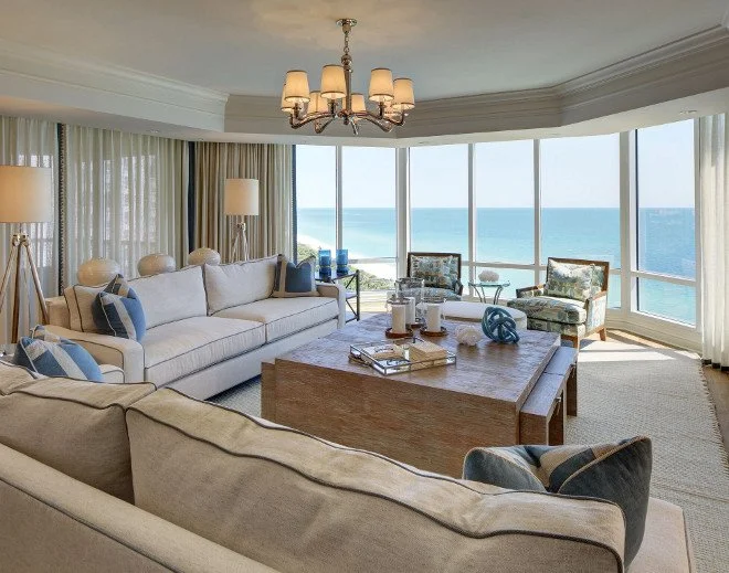 Living room with large windows overlooking the ocean, white sofas with blue and white pillows, two patterned armchairs, a wooden coffee table, and a chandelier.