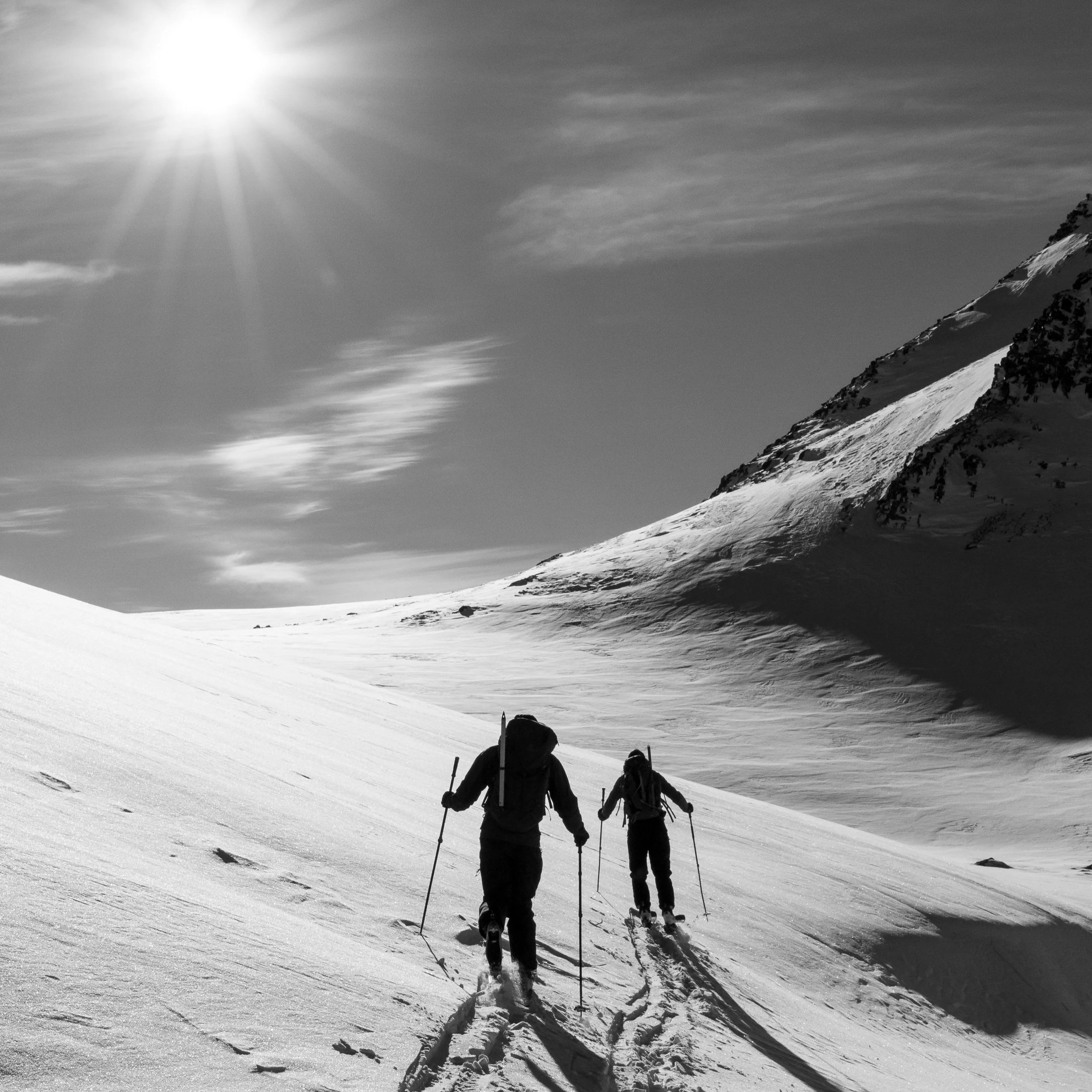 Two people hiking on a snow-covered mountain slope in bright sunlight, with one mountain peak on the right and a partly cloudy sky overhead.