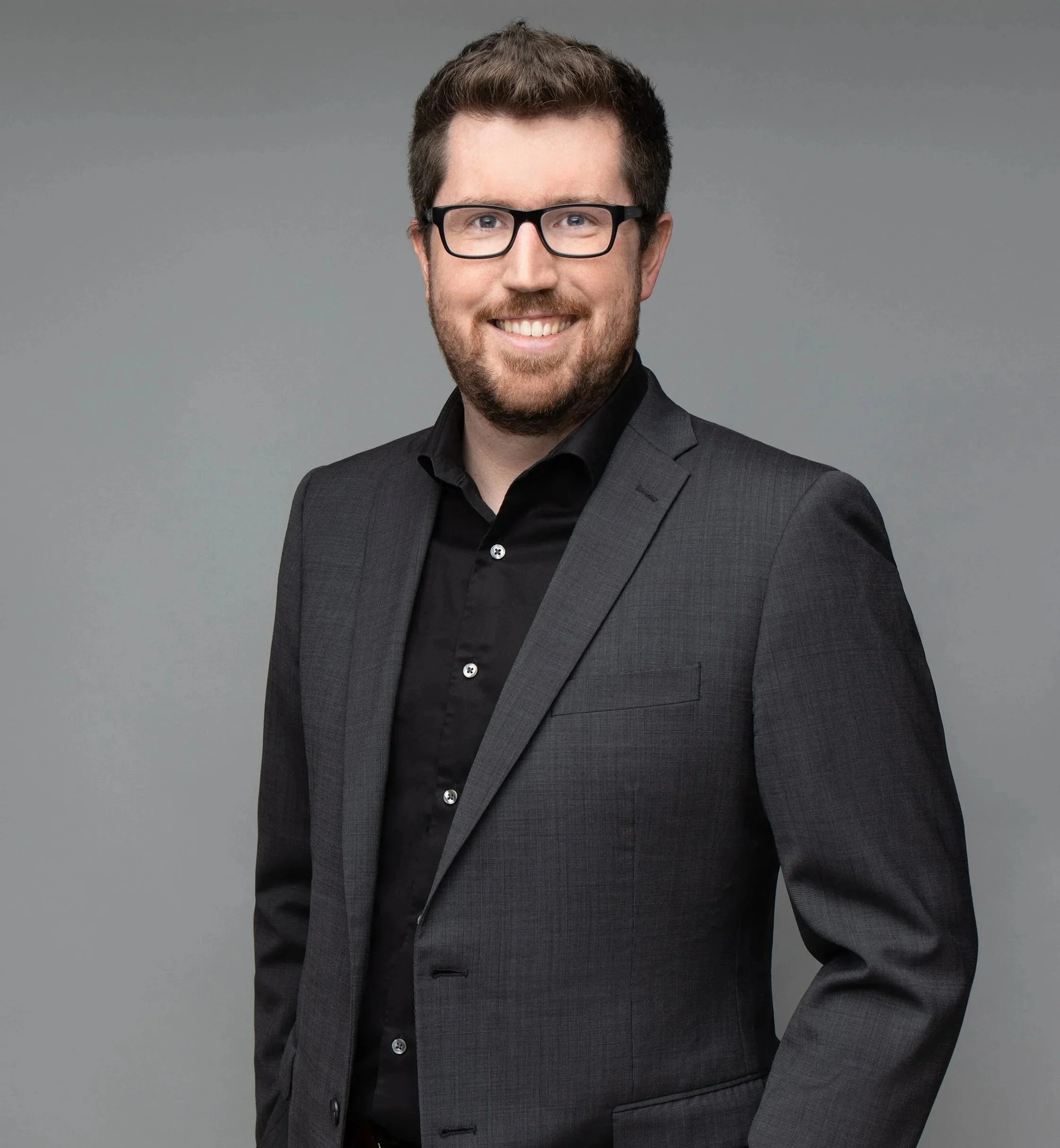 A man with glasses, a beard, and brown hair in a professional black suit and shirt, smiling against a gray background.
