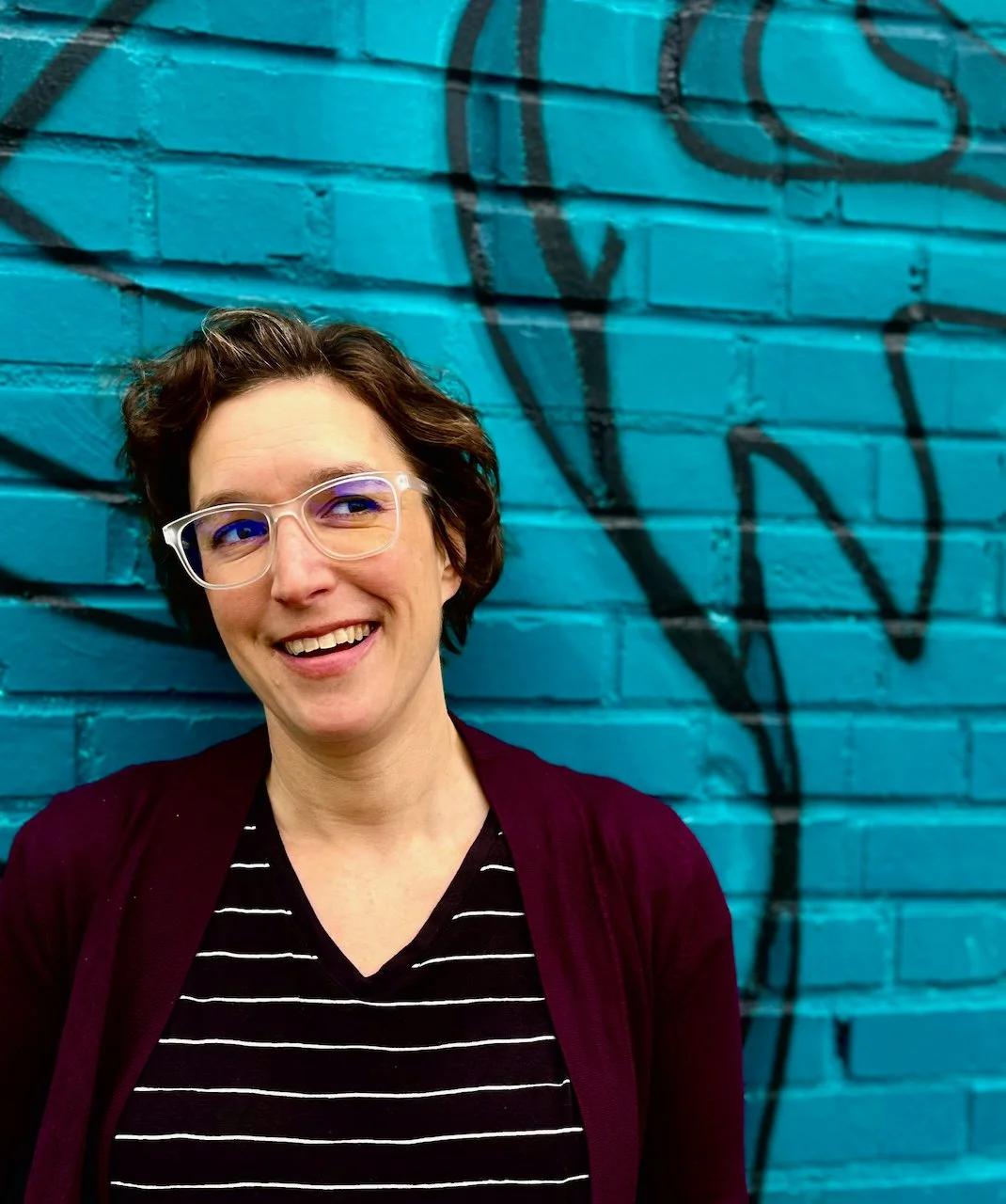 white woman with clear glasses and short brown hair in a black and white striped shirt in front of a blue wall with graffiti on it