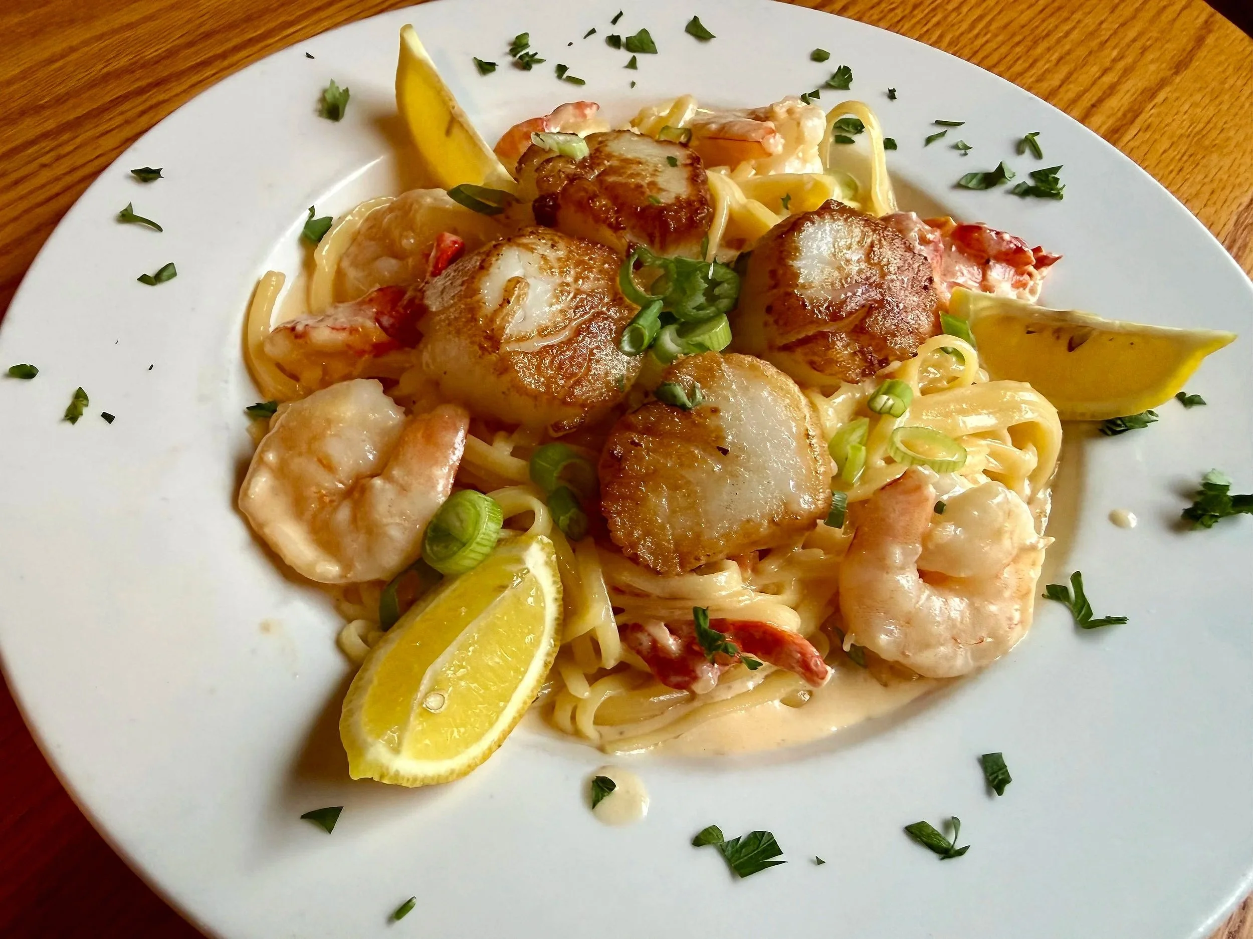 Linguine pasta with lobster, lemon wedges, chopped green onions, and fresh herbs on a white plate.
