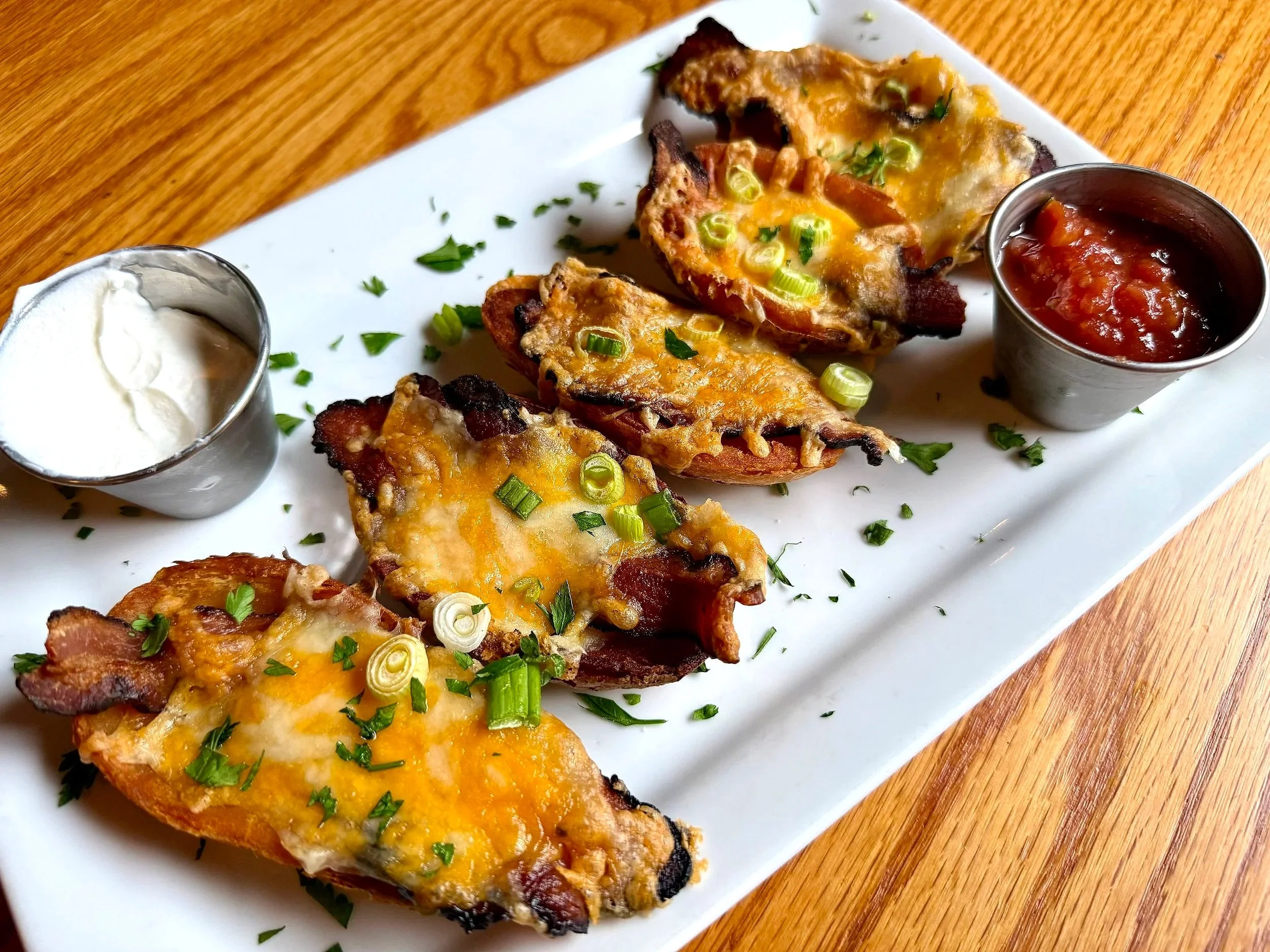 Baked potato skins topped with melted cheese, green onions, and chopped herbs, served with sour cream and salsa on a white rectangular plate.
