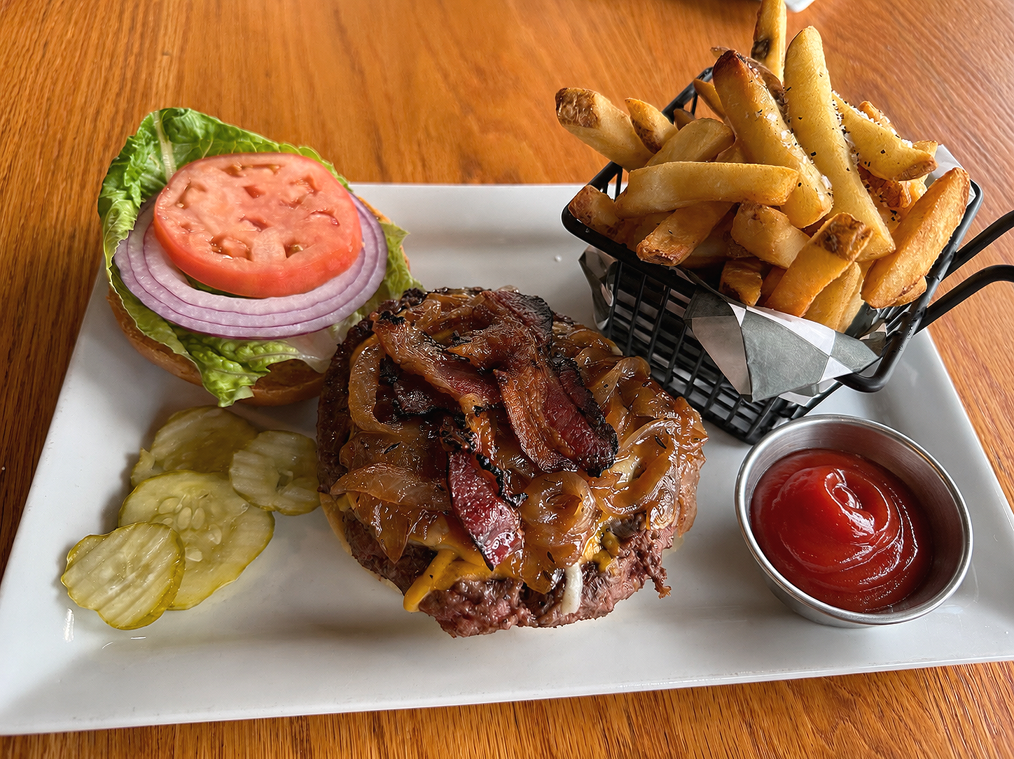 Burger with lettuce, tomato, onions, pickles, French fries with salt, and ketchup on a white rectangular plate on a wooden table.