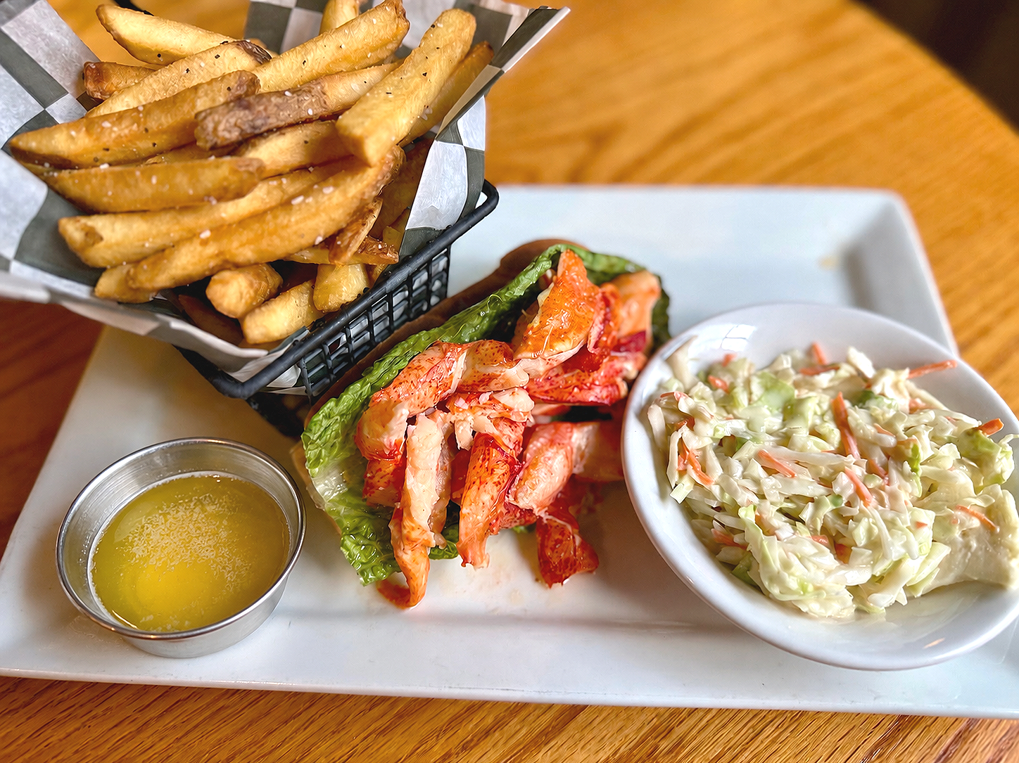 Basket of French fries with herbs, a bowl of coleslaw, a lobster roll with lettuce and lobster meat, and a small cup of melted butter on a white platter.
