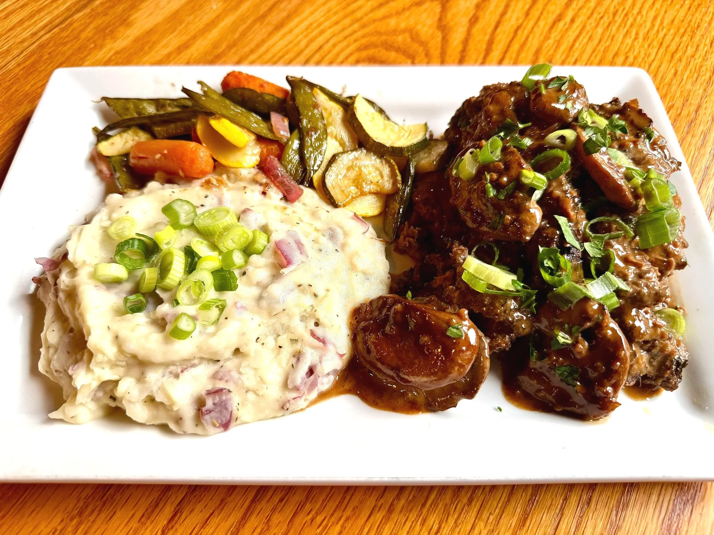 A white rectangular plate with a serving of potato salad sprinkled with chopped green onions, roasted vegetables including zucchini, carrots, and yellow squash, and pieces of beef stew garnished with chopped green onions on top, on a wooden table.