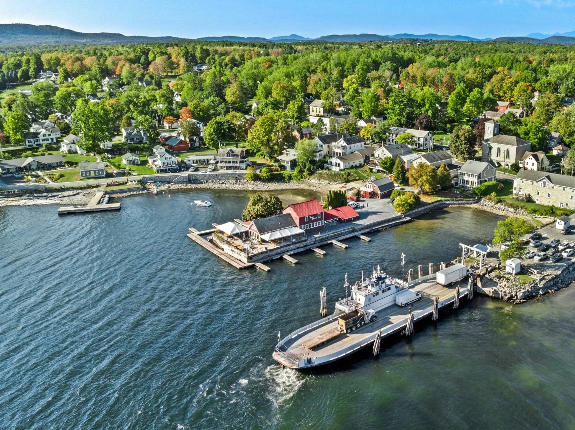 Aerial view of a waterfront town with boats docked at piers, green trees, and houses in the background under a clear sky.