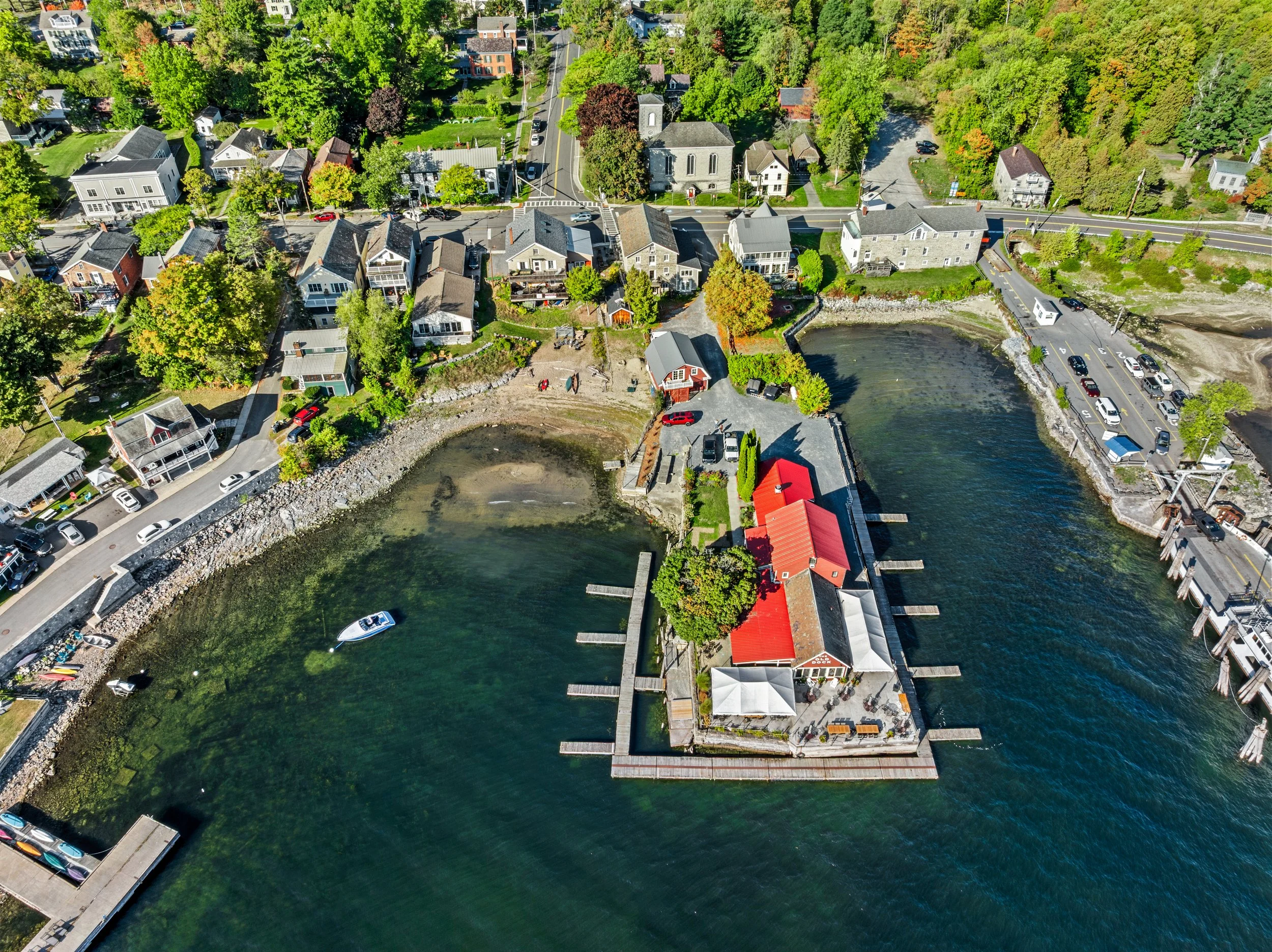 Aerial view of a coastal neighborhood with houses, trees, and water. There's a waterfront area with docks, boats, and a red-roofed building.