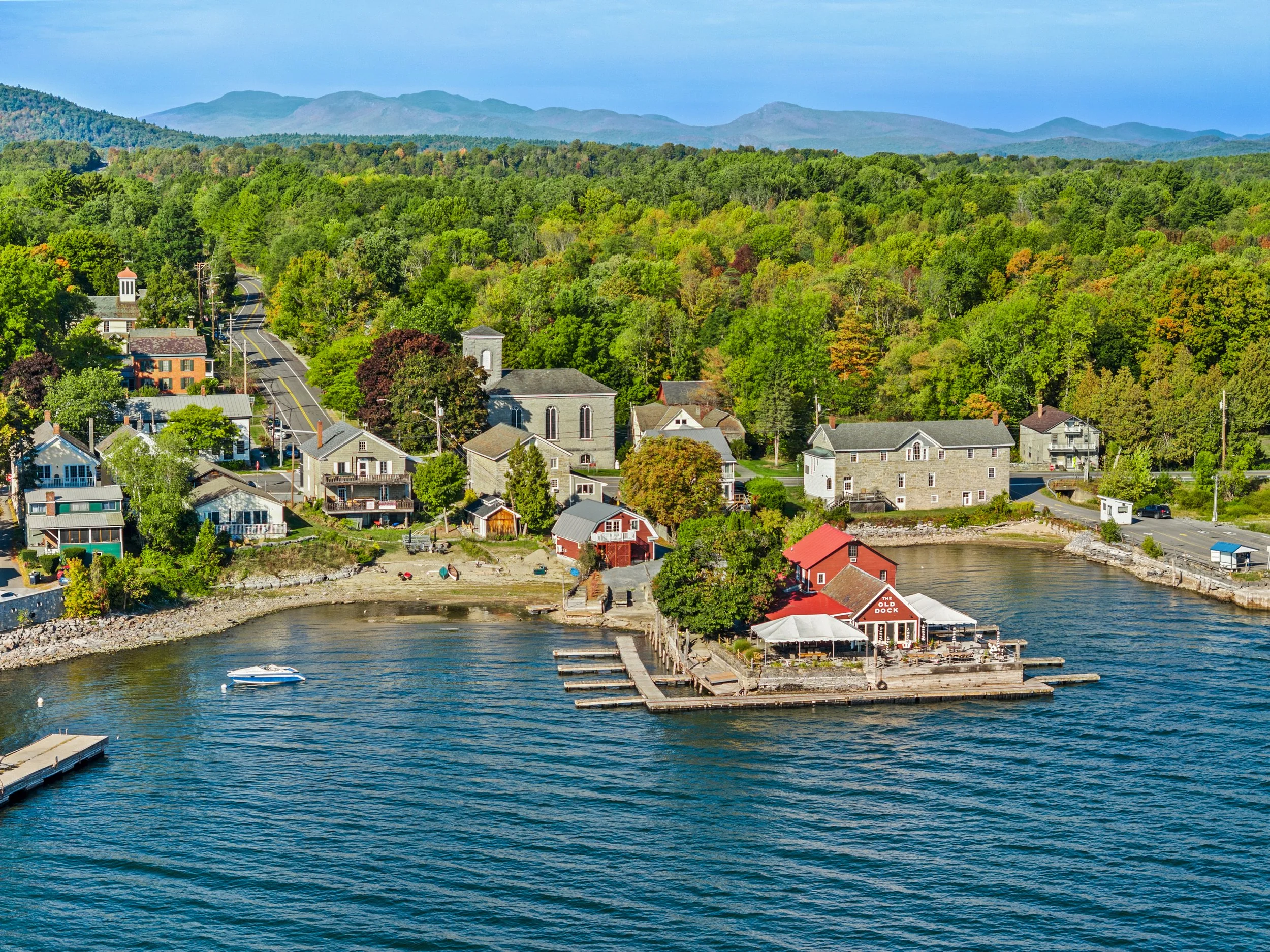 A small waterfront town with colorful houses, a church, and a dock, surrounded by lush green trees and mountains in the distance, under a blue sky.