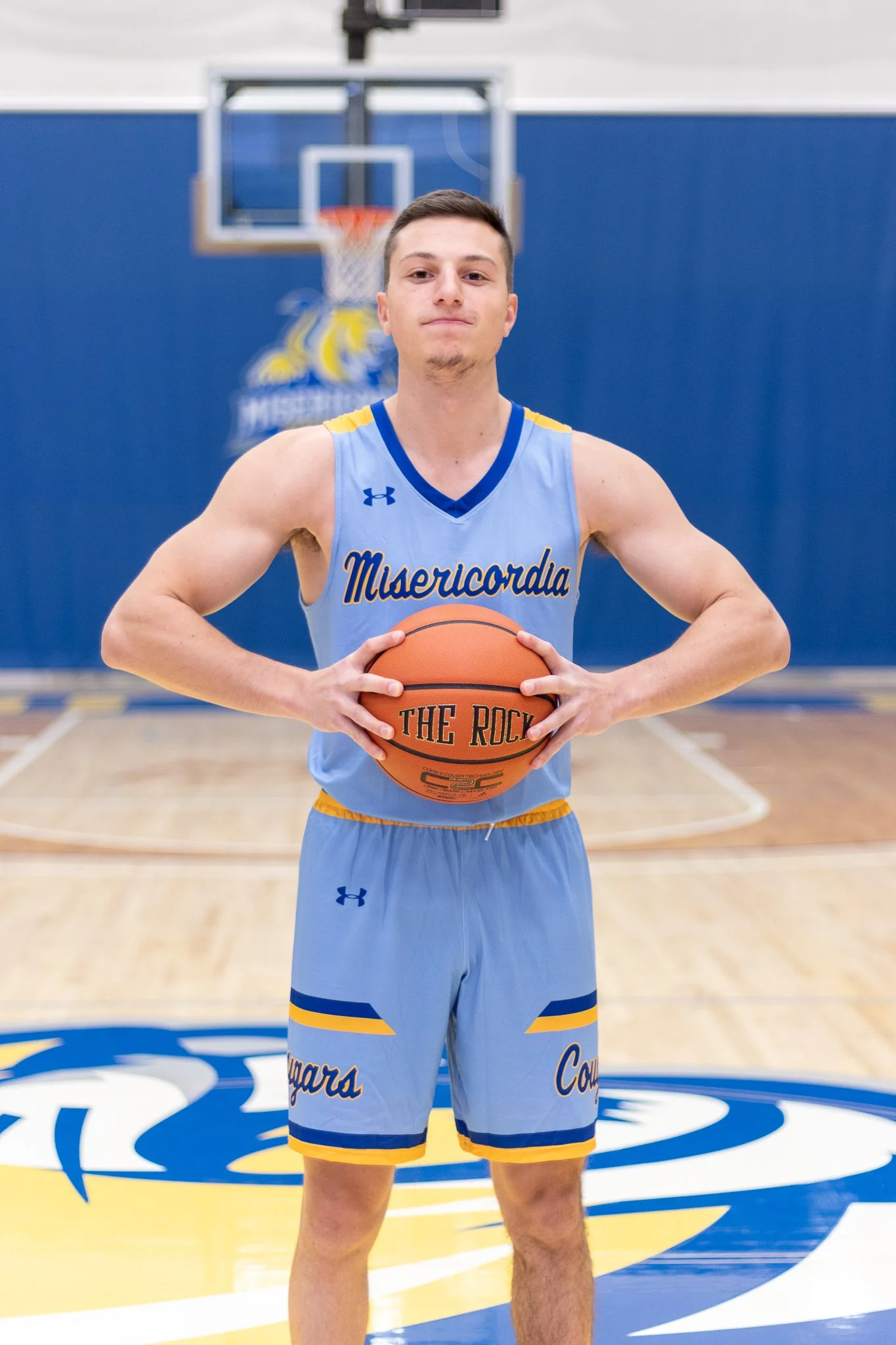 Young male basketball player in light blue and yellow uniform holding an orange basketball on a basketball court, standing in front of a hoop and blue wall with a logo.