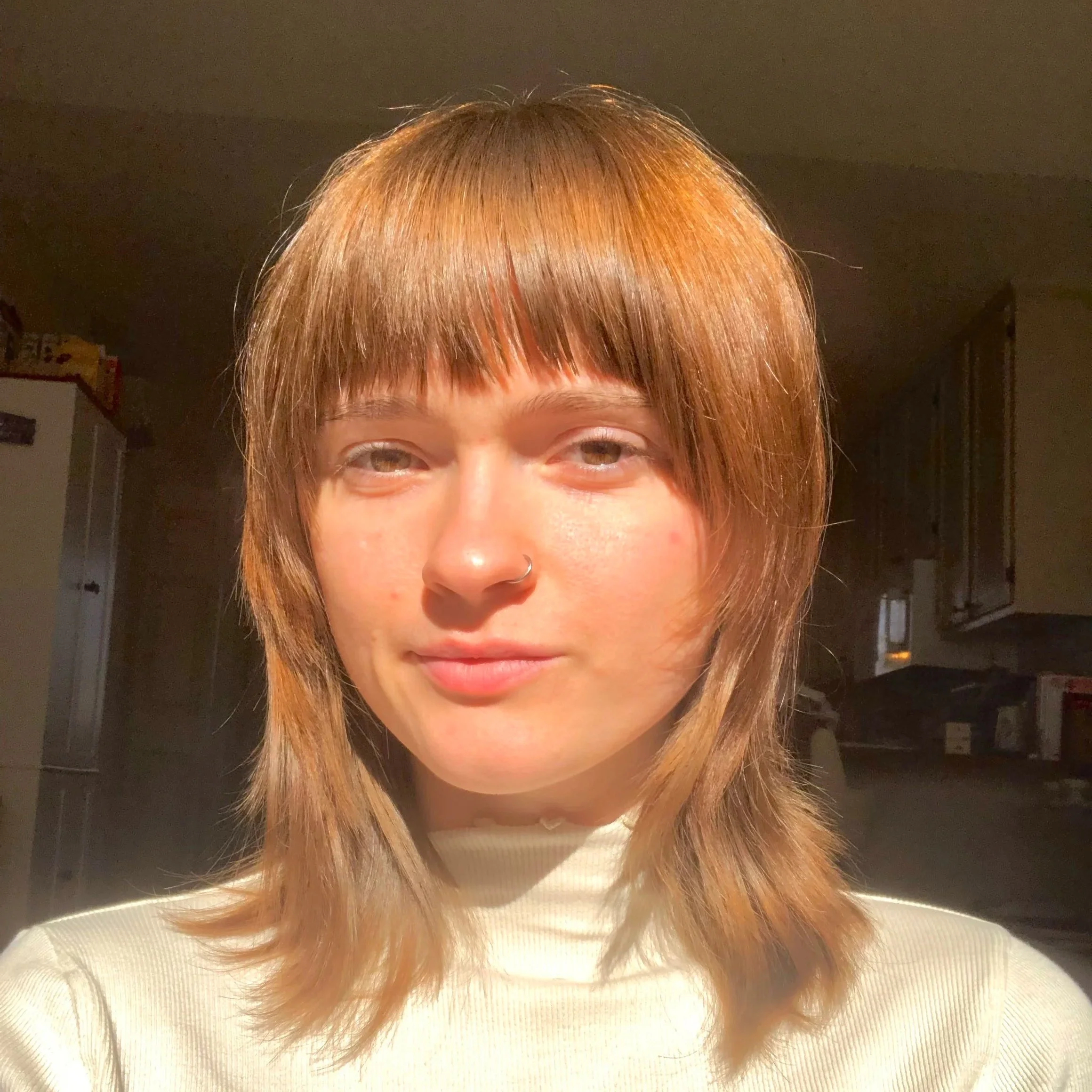Young woman with light brown hair and a nose ring, taking a selfie in a kitchen lit by warm sunlight.