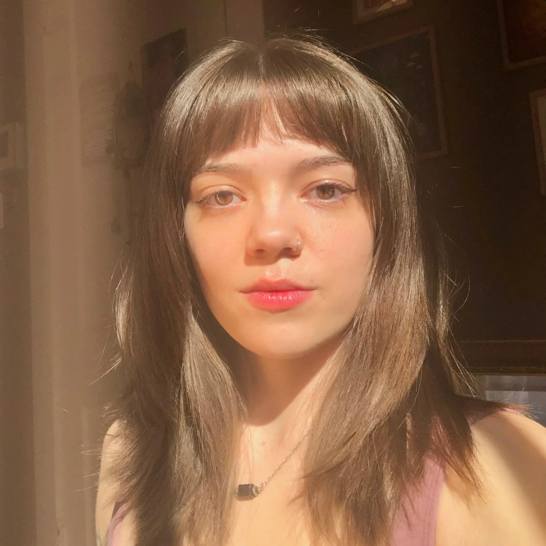 Close-up of a young woman with shoulder-length brown hair and a nose ring, looking at the camera with a neutral expression in warm indoor lighting.