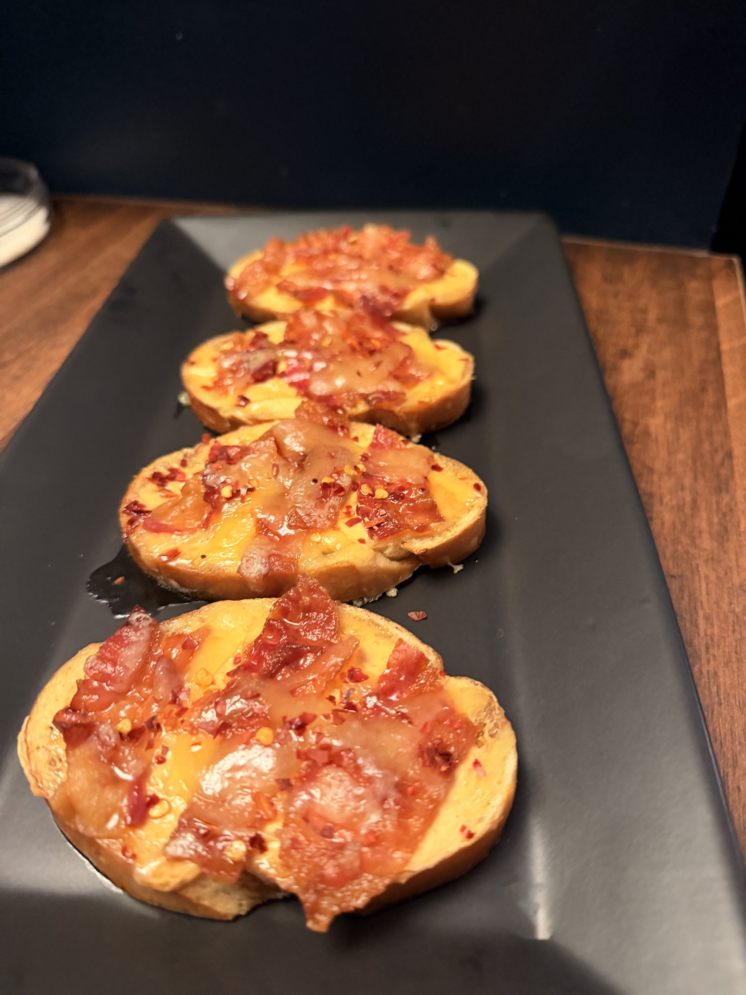 Four slices of bread topped with melted cheese, diced tomatoes, bacon bits, and red pepper flakes, served on a black rectangular plate.