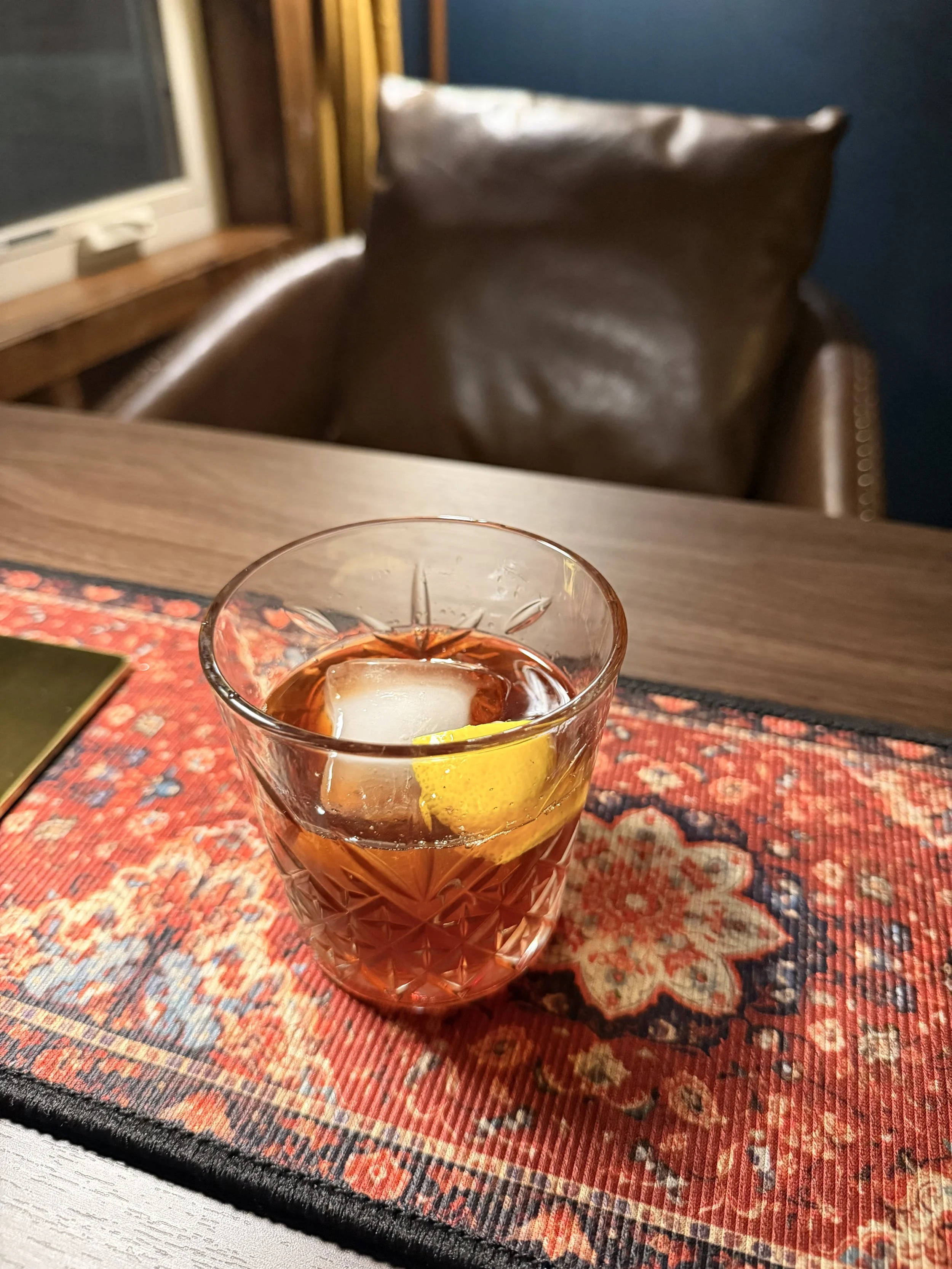Close-up of a glass of iced tea with a lemon slice and ice cubes on a decorative tablecloth, with a brown leather armchair, blue walls, and a window in the background.