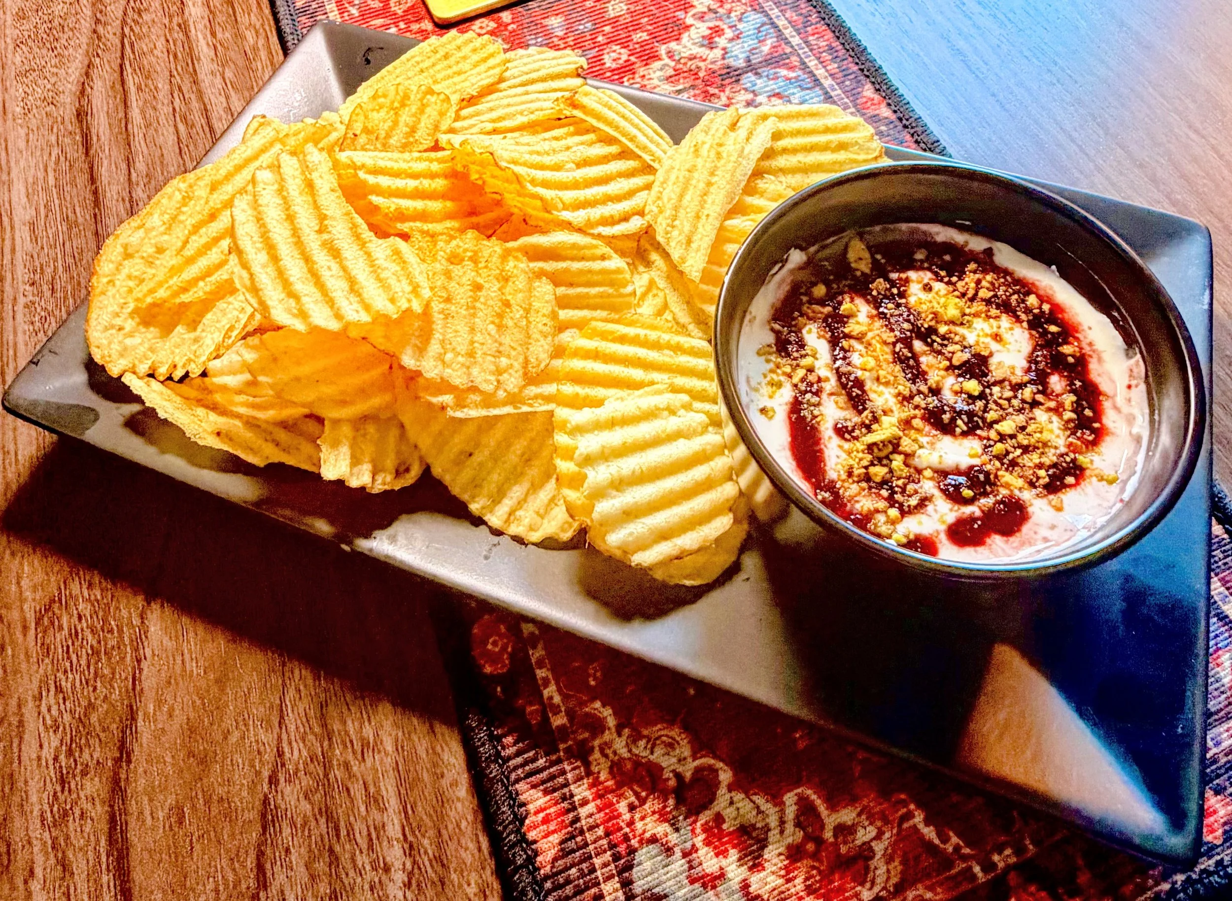 A serving of ridged potato chips in a rectangular dish next to a bowl of creamy dip topped with red sauce, crushed nuts, and yellow seasoning, placed on a wooden table.