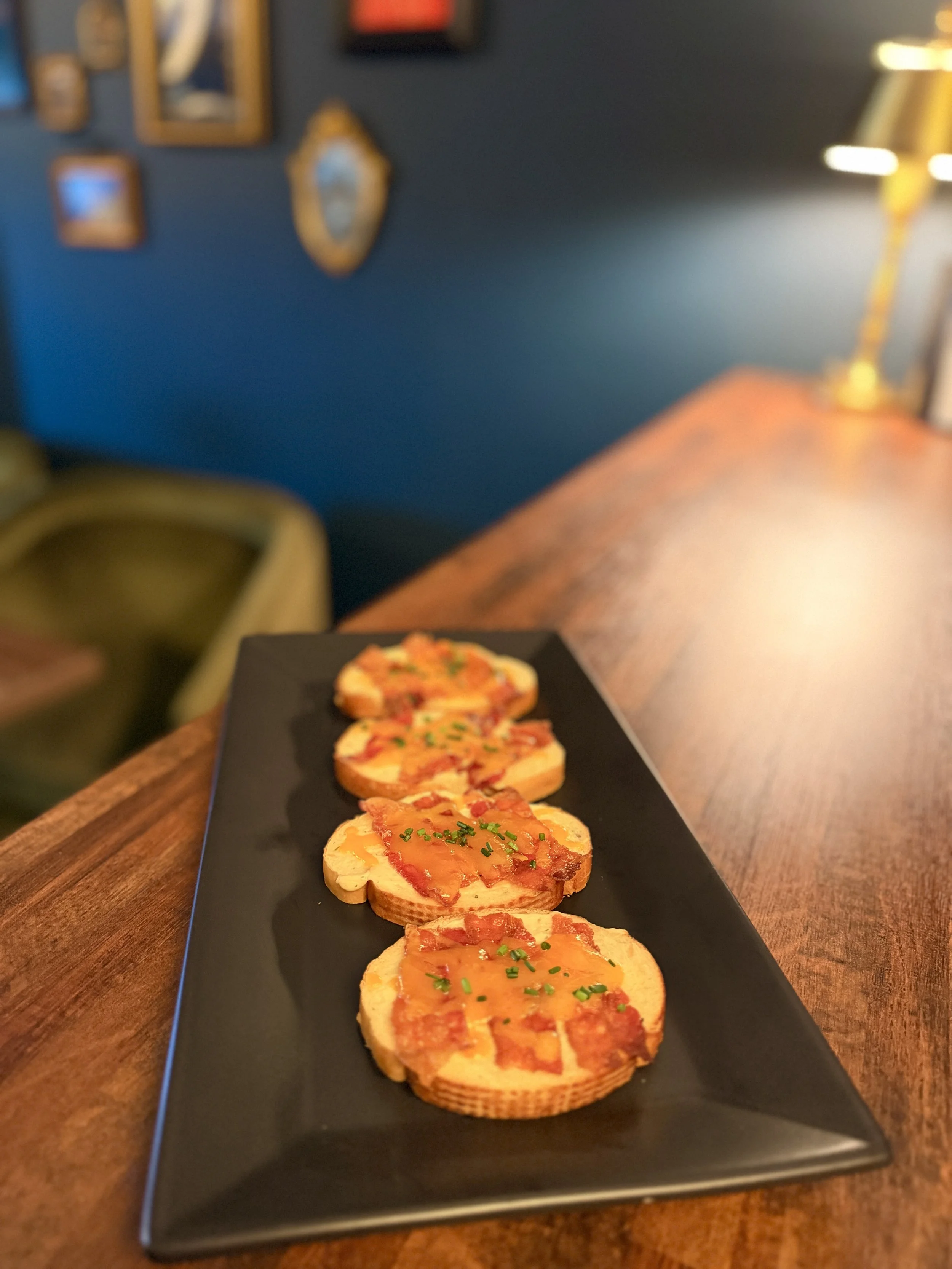 Close-up of five crostini appetizers with cheese, tomato, and green herbs on a black rectangular plate.