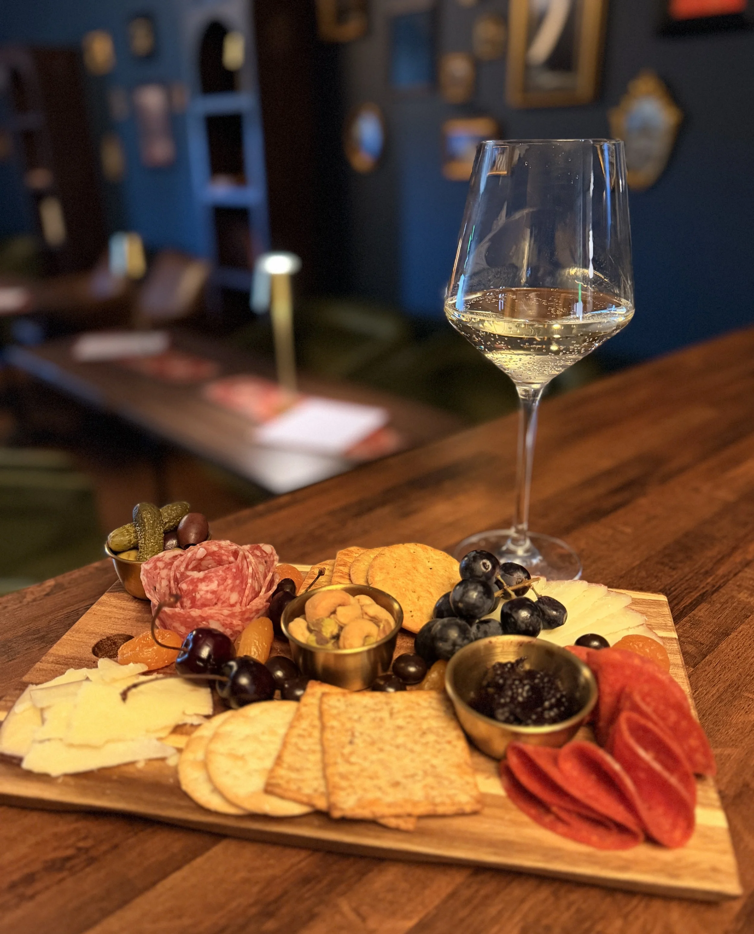 A cheese and charcuterie board with various cheeses, cured meats, grapes, olives, crackers, and small bowls of nuts and dried fruits on a wooden serving board, with a glass of white wine in the background.