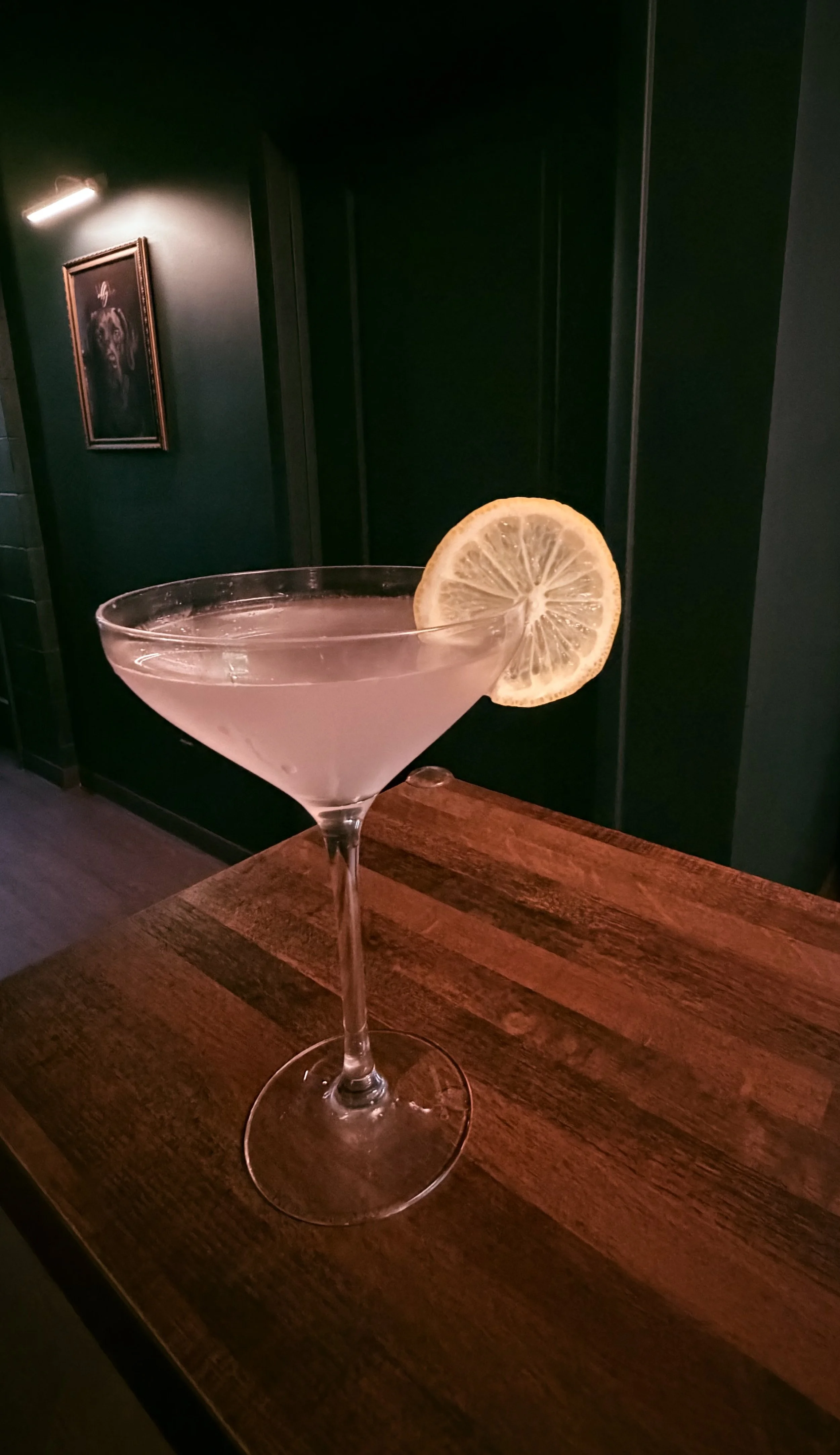 A cocktail glass with a lemon slice on the rim, placed on a wooden table in a dimly lit bar or restaurant.