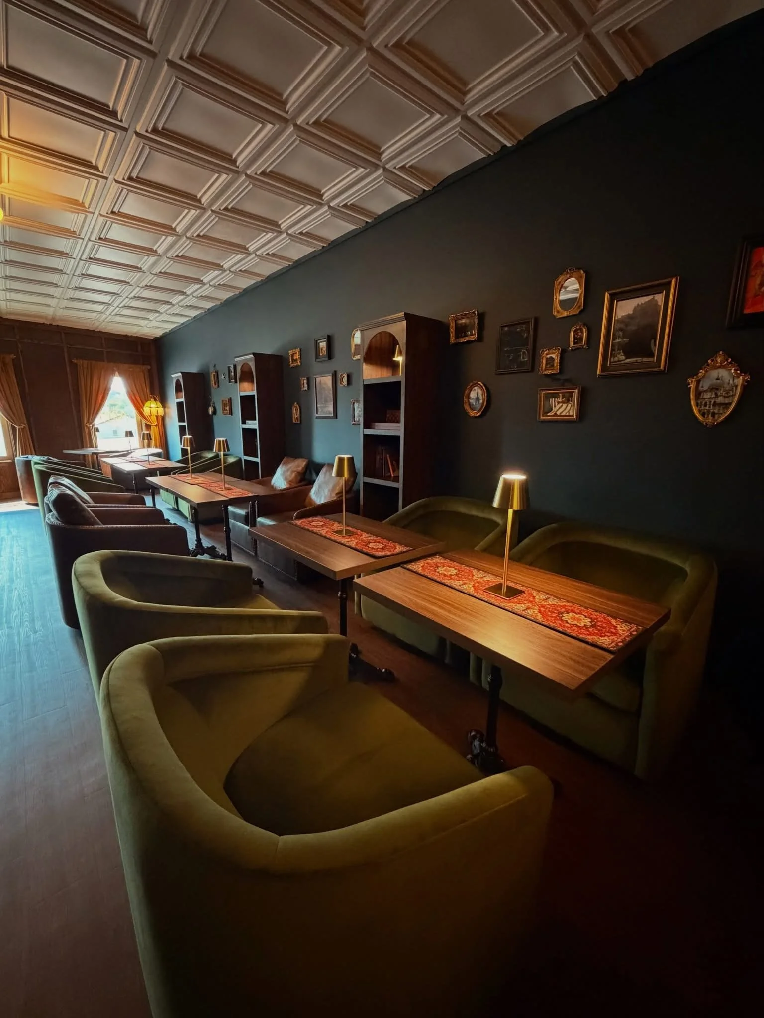 Elegant lounge with green velvet sofas, wooden tables with decorative runners, vintage picture frames on dark-colored wall, large window with curtains, and decorative lamps.