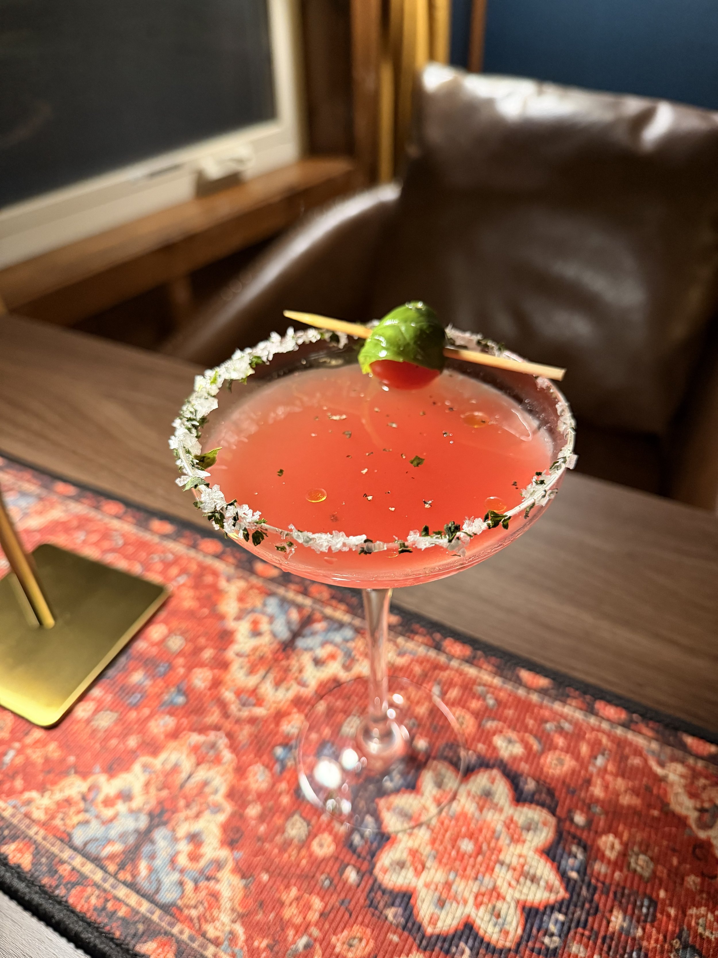 A pink cocktail in a margarita glass with a salted rim, garnished with a green olive on a skewer, sitting on a red and blue patterned rug.