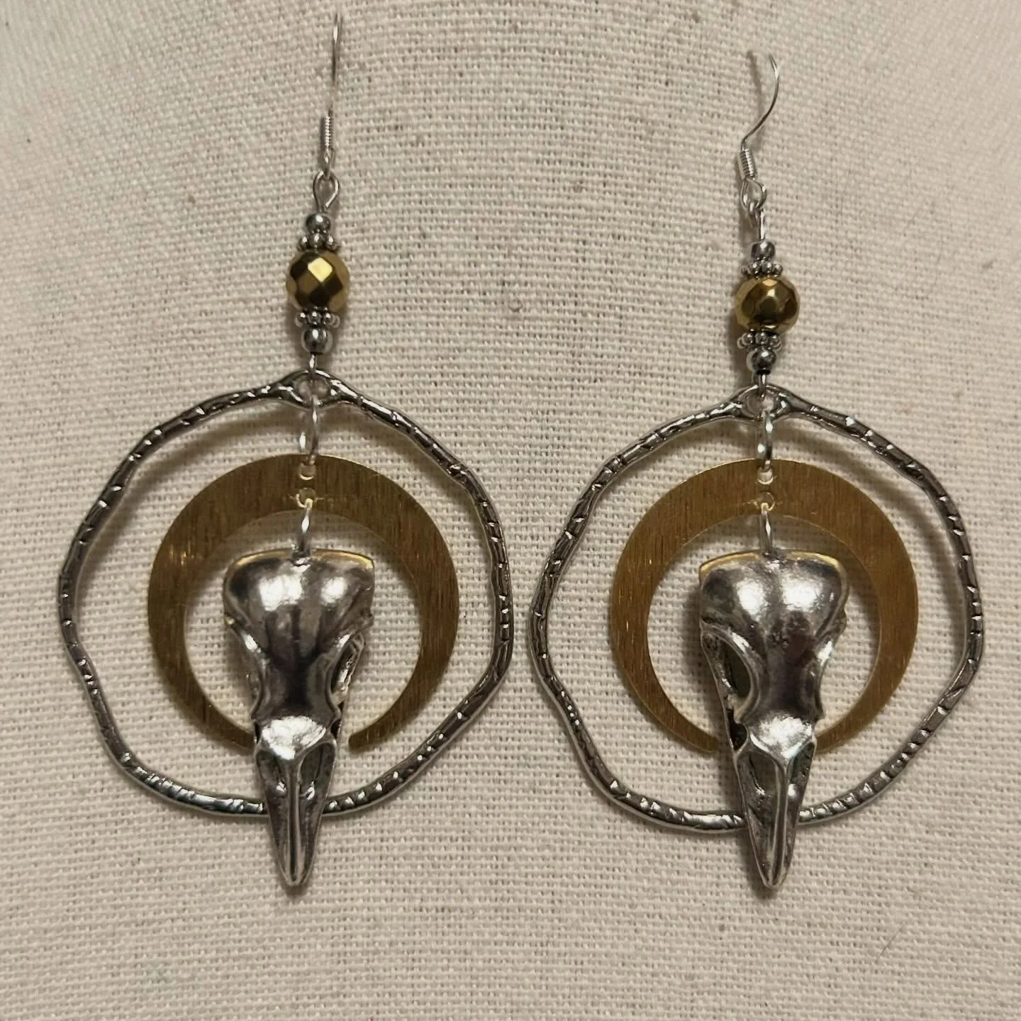 These statement earrings feature silver-tone raven skull charms suspended within layered silver and brass rings. They are accented with metallic faceted beads.